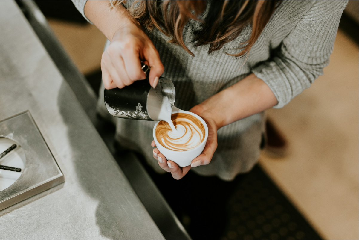 Barista som häller skummad mjölk i kaffe och skapar latte art i en kopp vid en cafédisk.