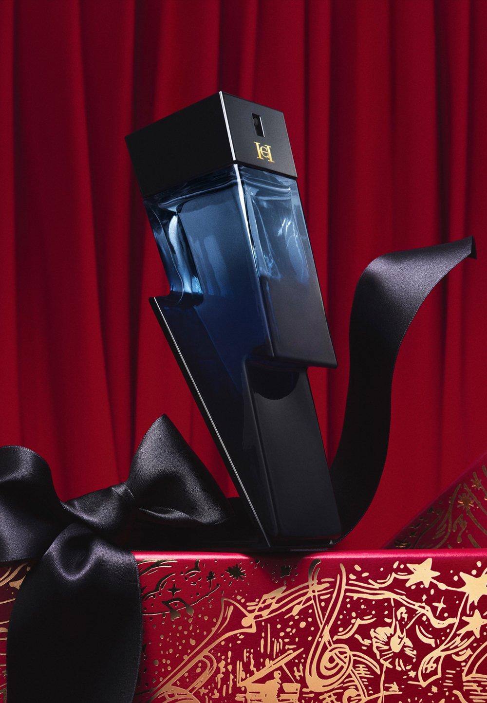 Blå parfymflaska formad som en blixt står på ett röd-guldigt bord med svart sidenrosett.