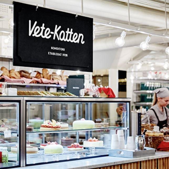 Skylt för konditori Vete-Katten ovanför disk med bakverk och personal i cafémiljö.