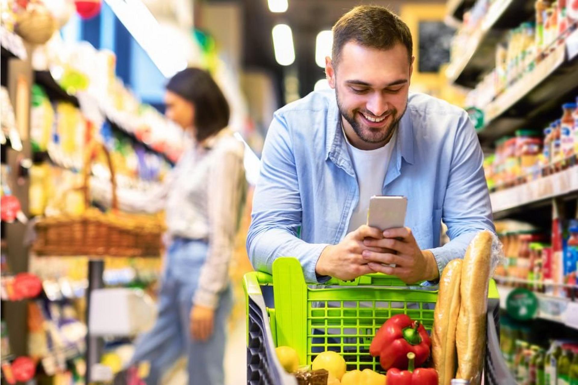 Man using smartphone while leaning on grocery cart with baguettes and produce.