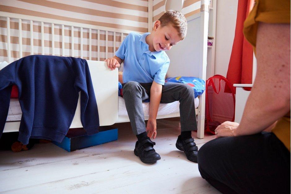 Child in blue shirt sitting on bed, reaching for shoe. Adult kneeling nearby. Sweater on bed.