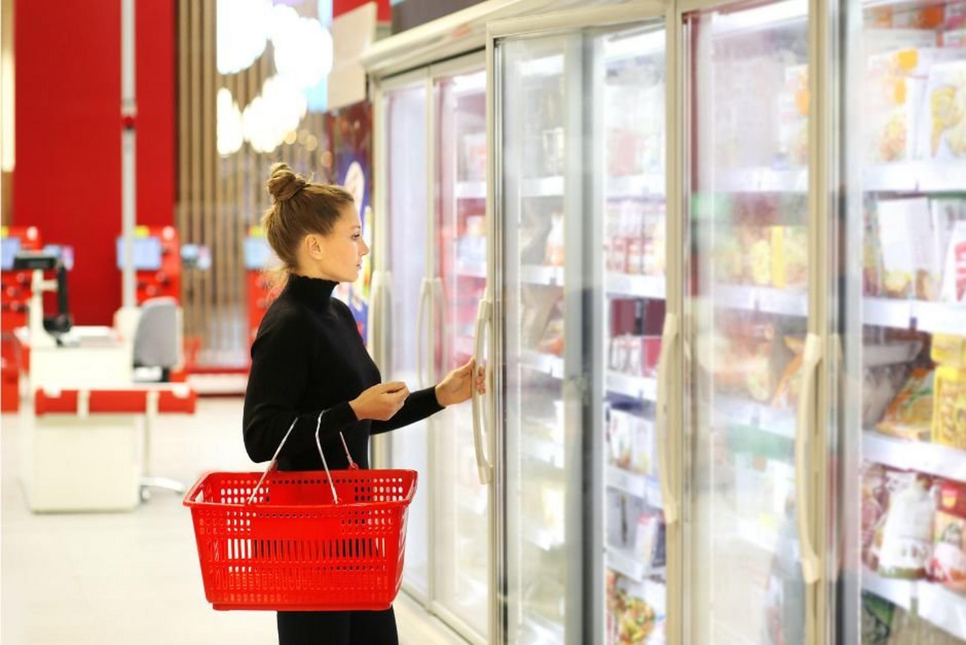Woman holding a shopping basket while opening a freezer door in a grocery store.