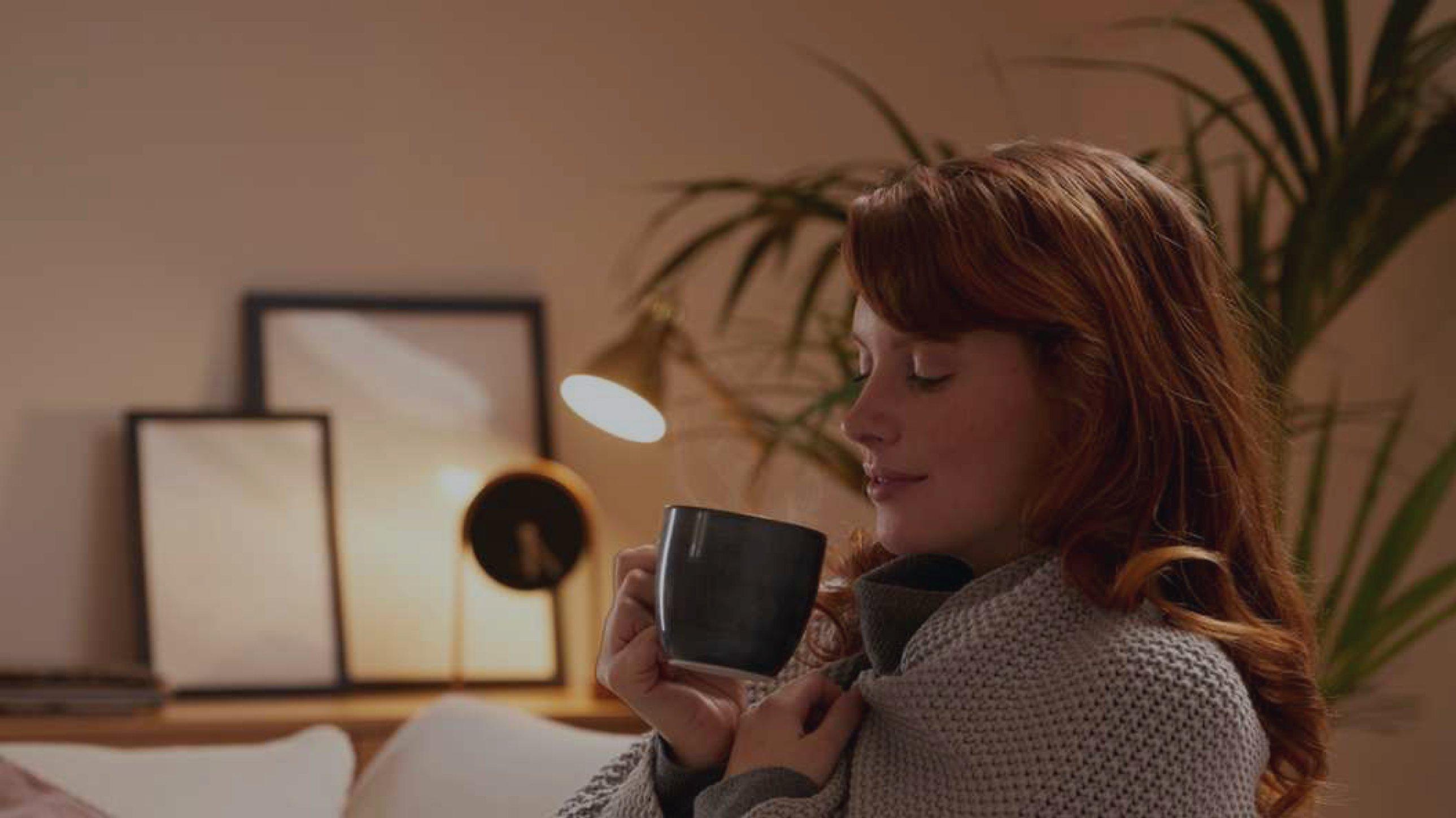 Woman sat on a sofa in a dimly lit room, wrapped in a blanket, about to take a drink from a black cup in her hand. A plant and picture frames are visible in the background.