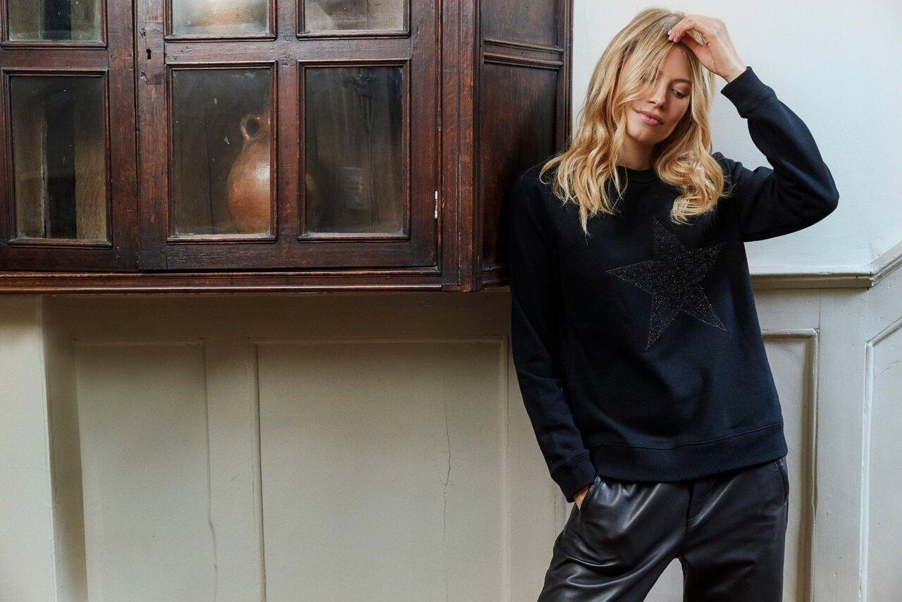 Woman in a black star sweater and leather pants, leaning against a wall with a wooden cabinet.