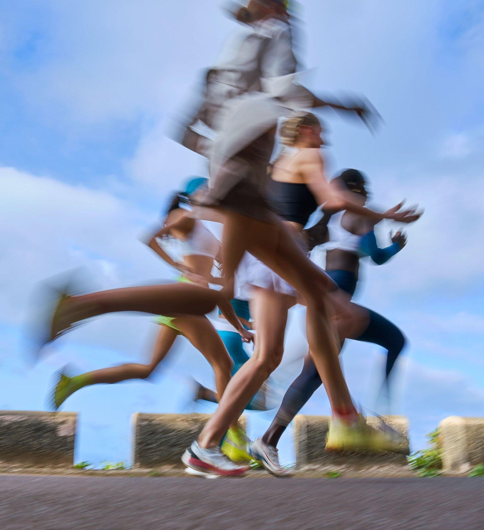 Blurred motion photo of people running outdoors on a paved path, with a clear sky in the background.
