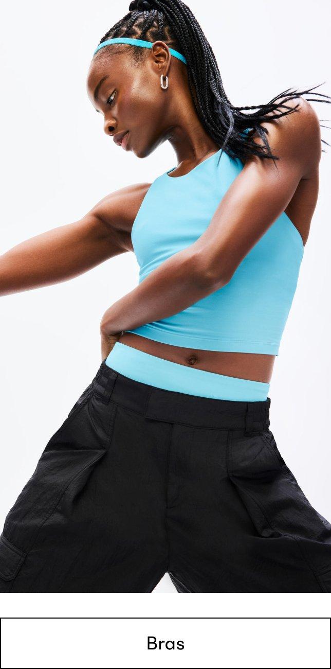 a woman in a aqua top, black pants, and aqua biker shorts layered underneath the black pants is posing. Shop Bras.