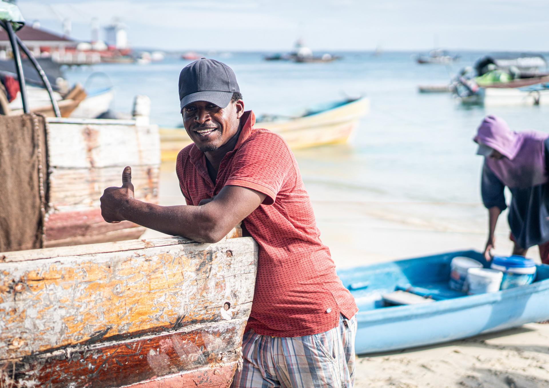 Man smiling and giving a thumbs-up by a weathered boat on a sunny beach with other boats and people in the background