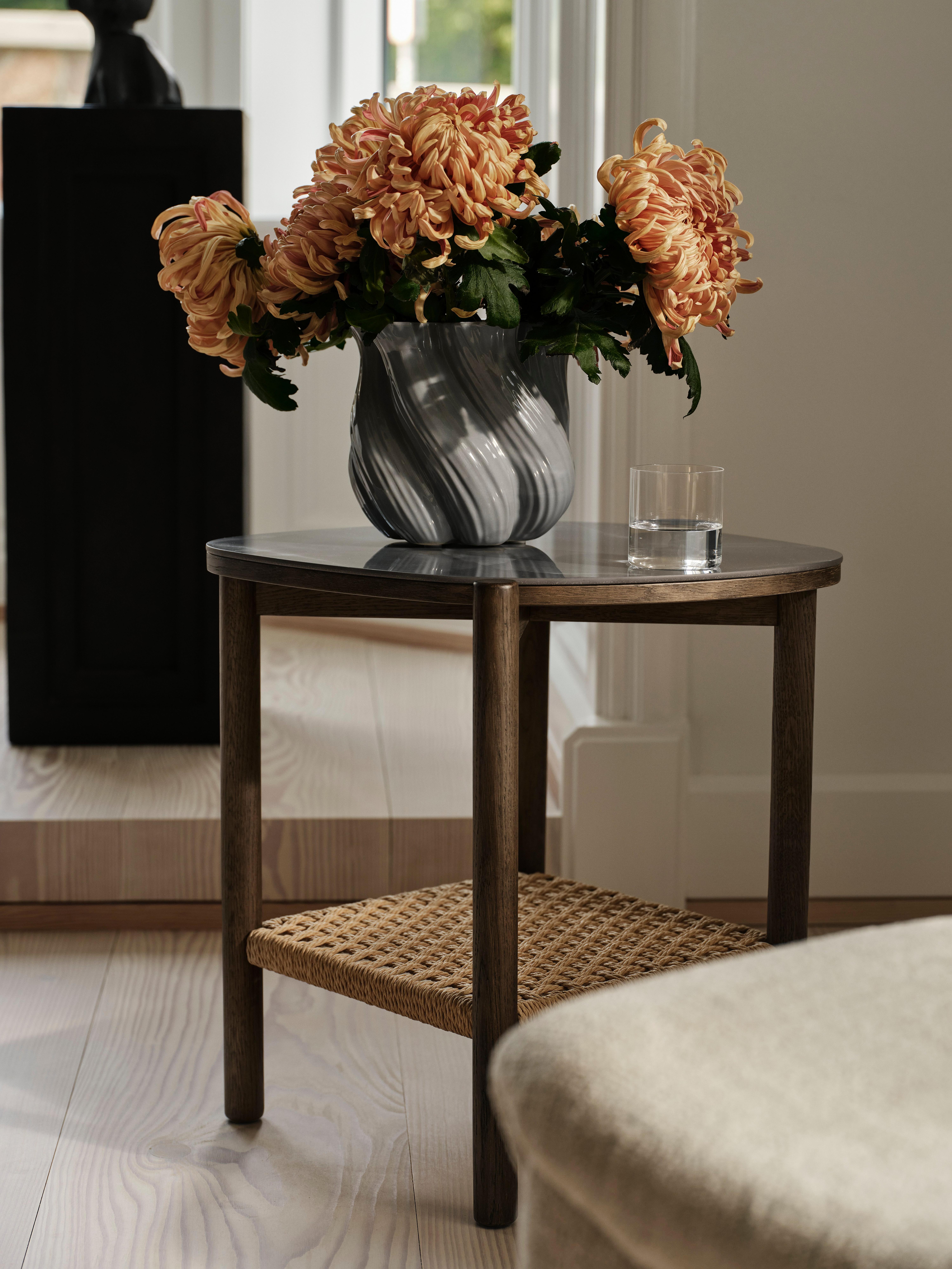 Vase mit orangefarbenen Blumen auf einem kleinen Holztisch, daneben ein Glas Wasser.