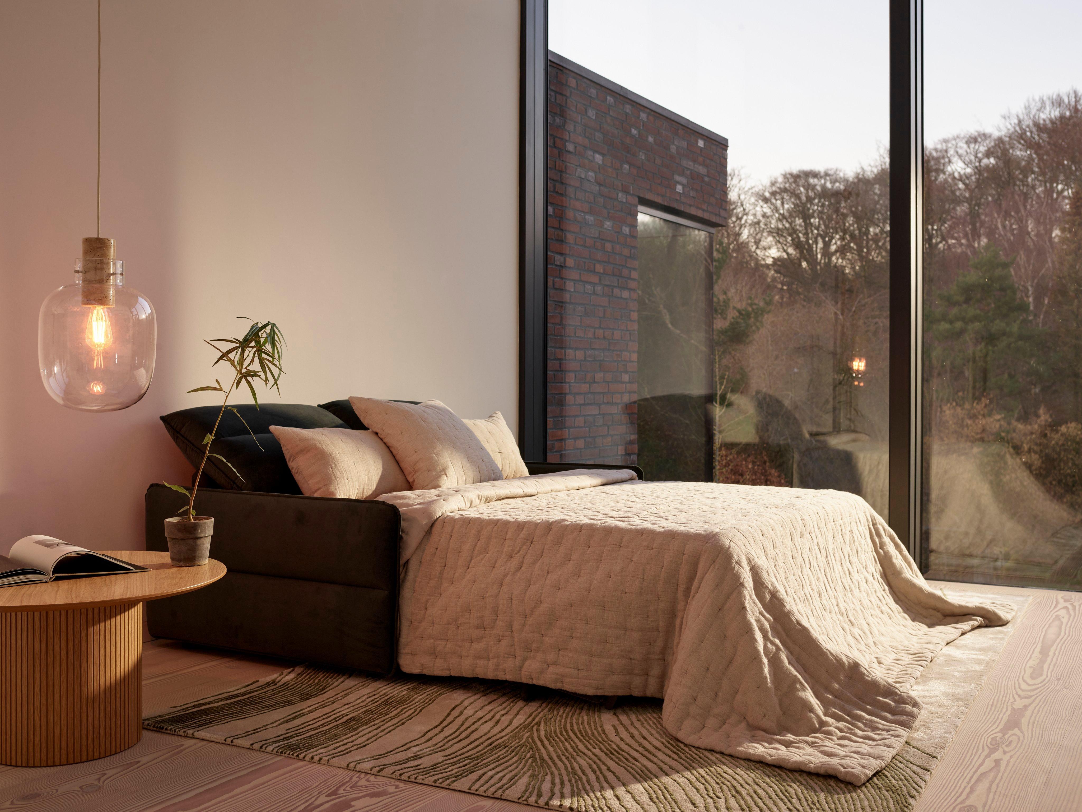 Modern living room converted to a bedroom with a dark green Southampton sofa bed, beige quilt, wooden Santiago table, and pendant light, by a floor-to-ceiling window.