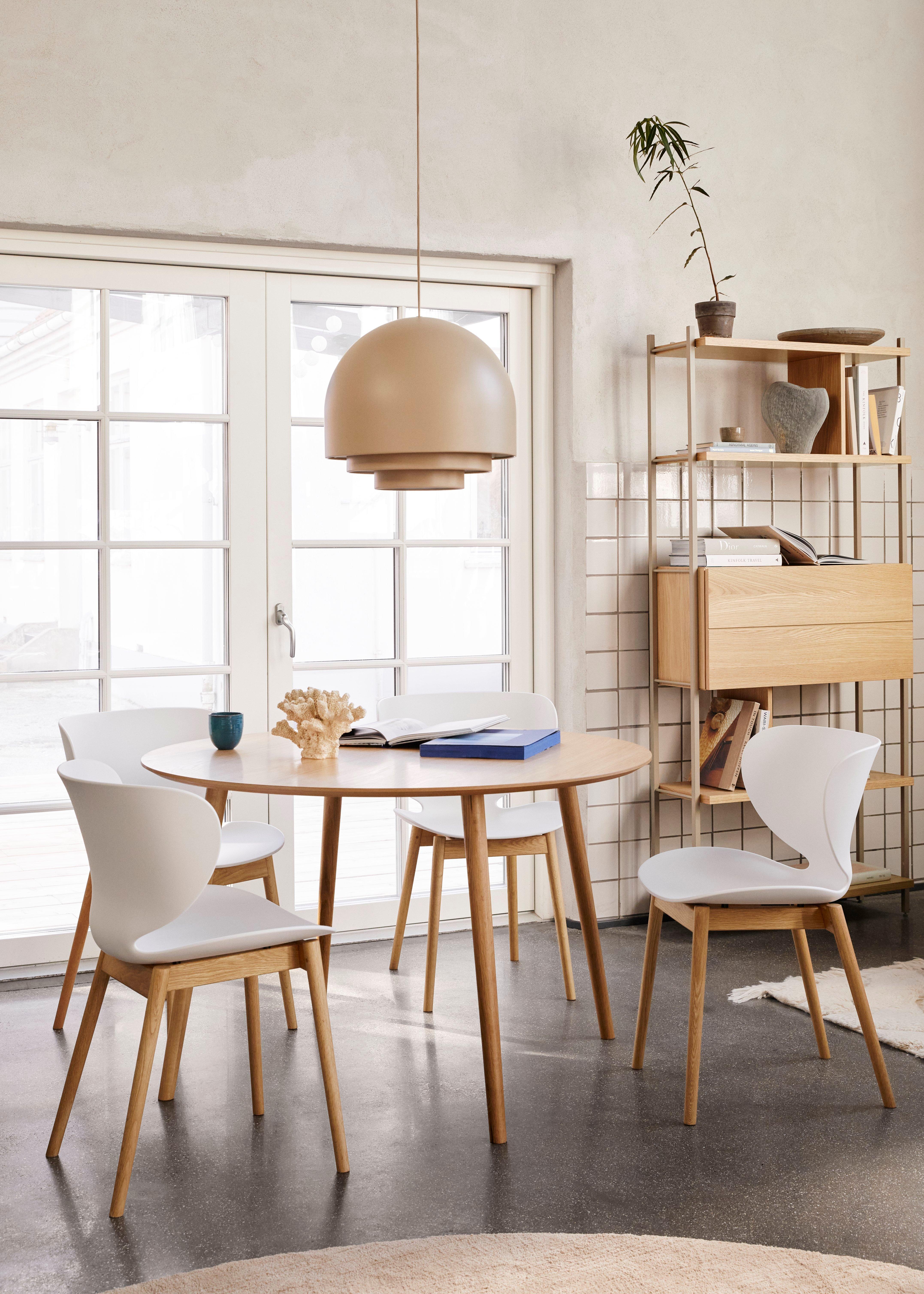 Round Bornholm dining table with four white Hamilton chairs under a beige pendant light, near a bookshelf and glass doors, in a modern interior.