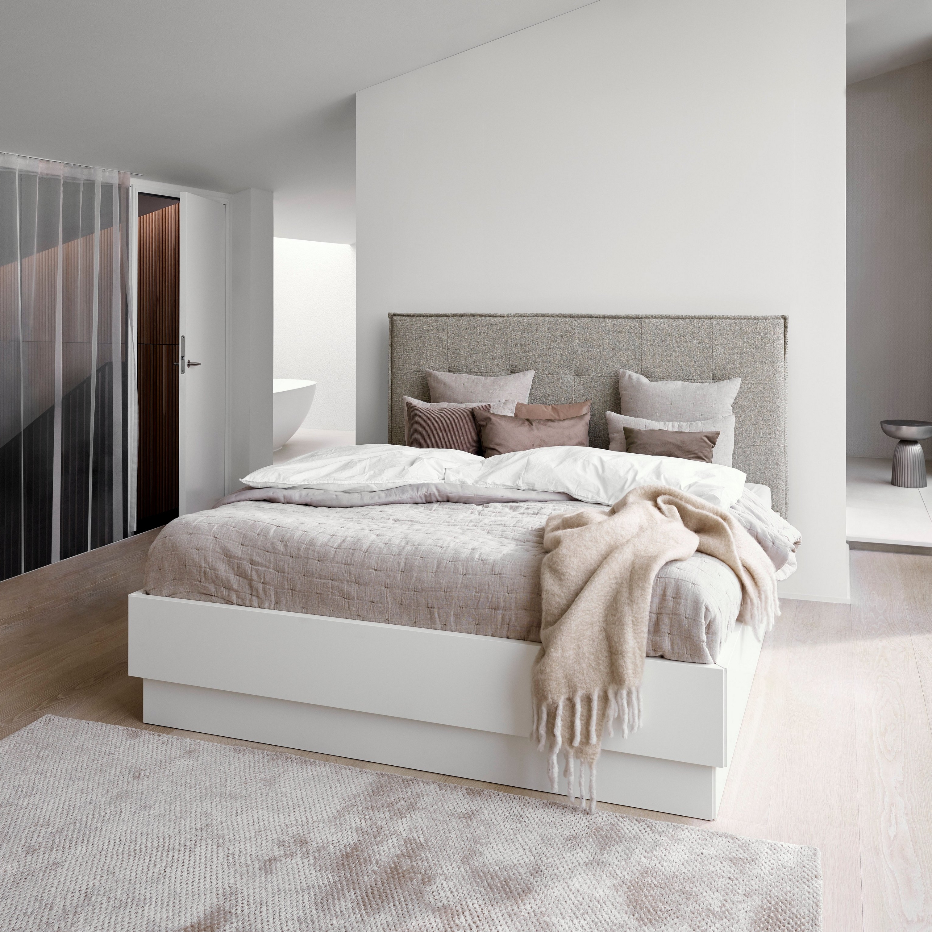 Dormitorio minimalista con cama tapizada gris, ropa de cama blanca y manta beige texturizada.