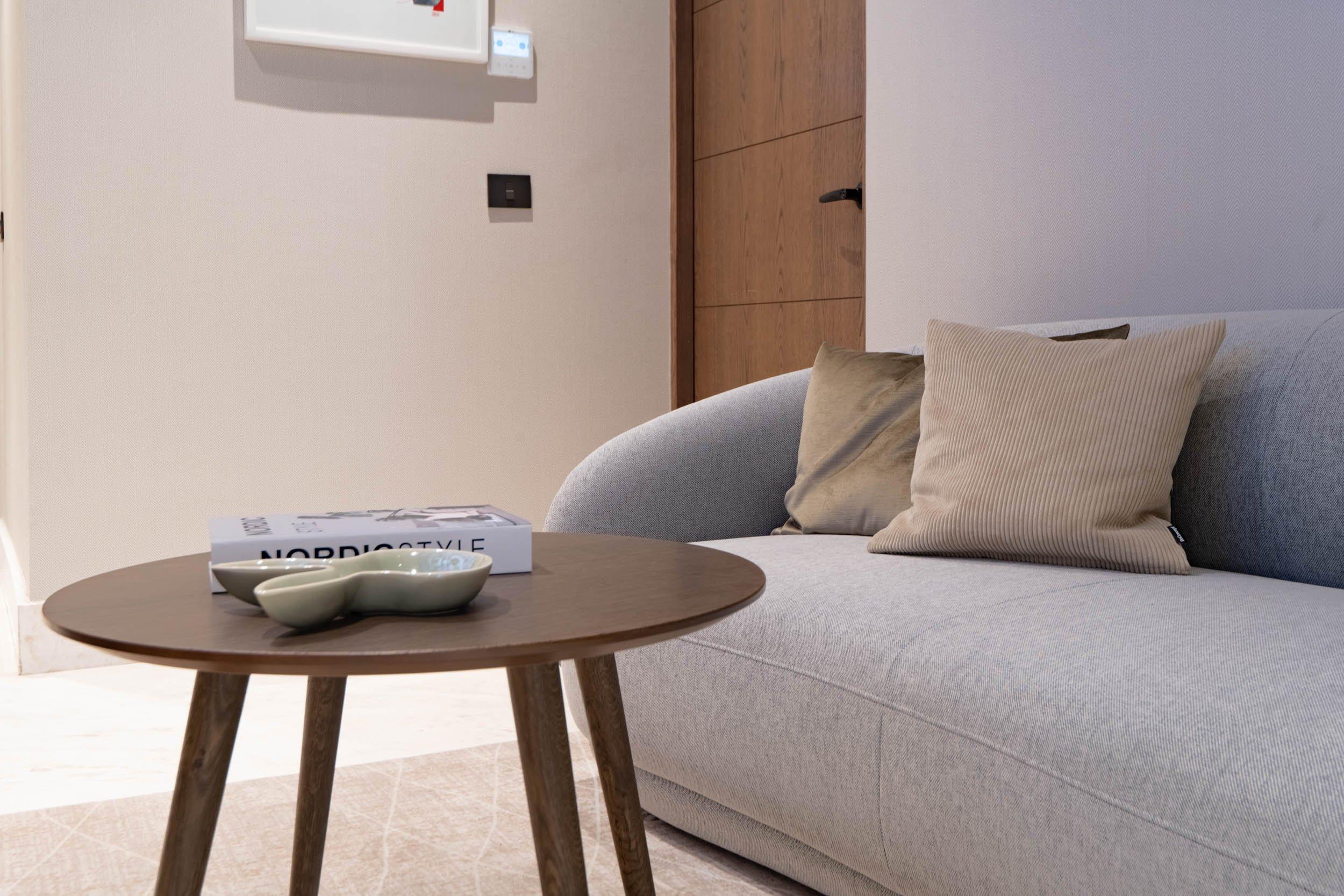 Table basse ronde en bois avec vide-poches, devant un canapé gris avec coussins.