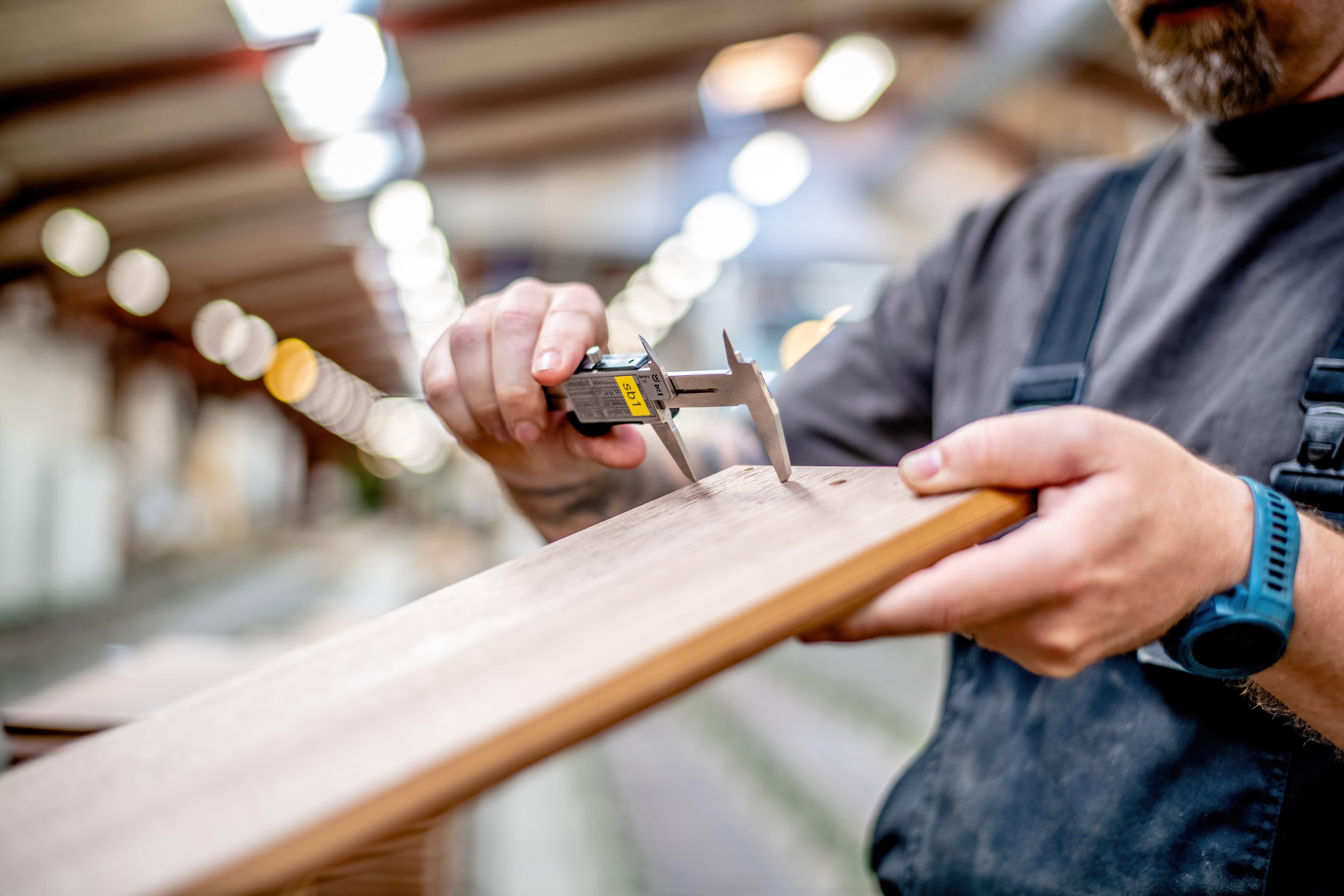 Un trabajador utiliza calibradores para medir el espesor de una tabla de madera en un taller bien iluminado con un fondo difuminado.