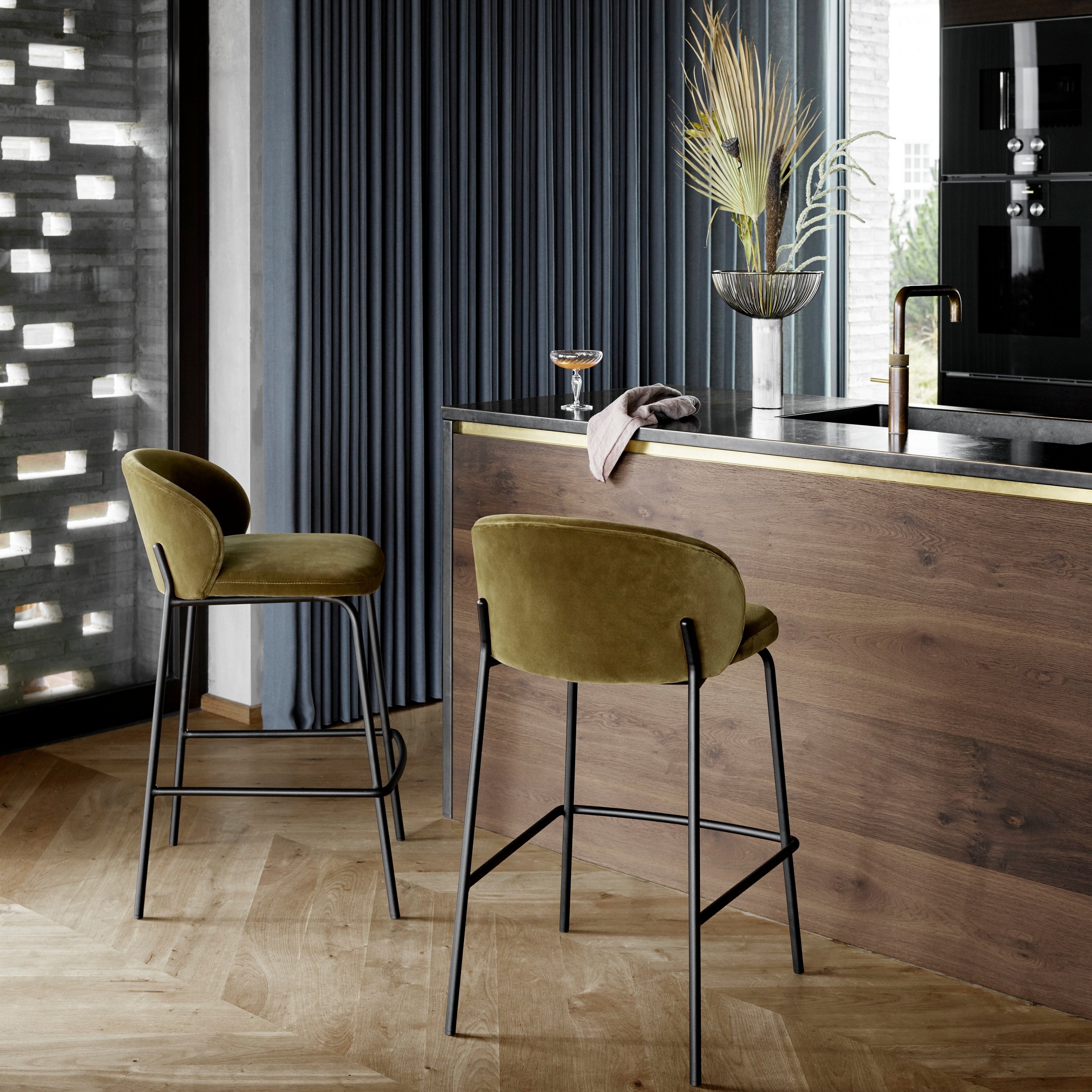 Barové stoličky v látke Velvet pri drevenom kuchynskom pulte s čiernymi spotrebičmi a závesným svietidlom.