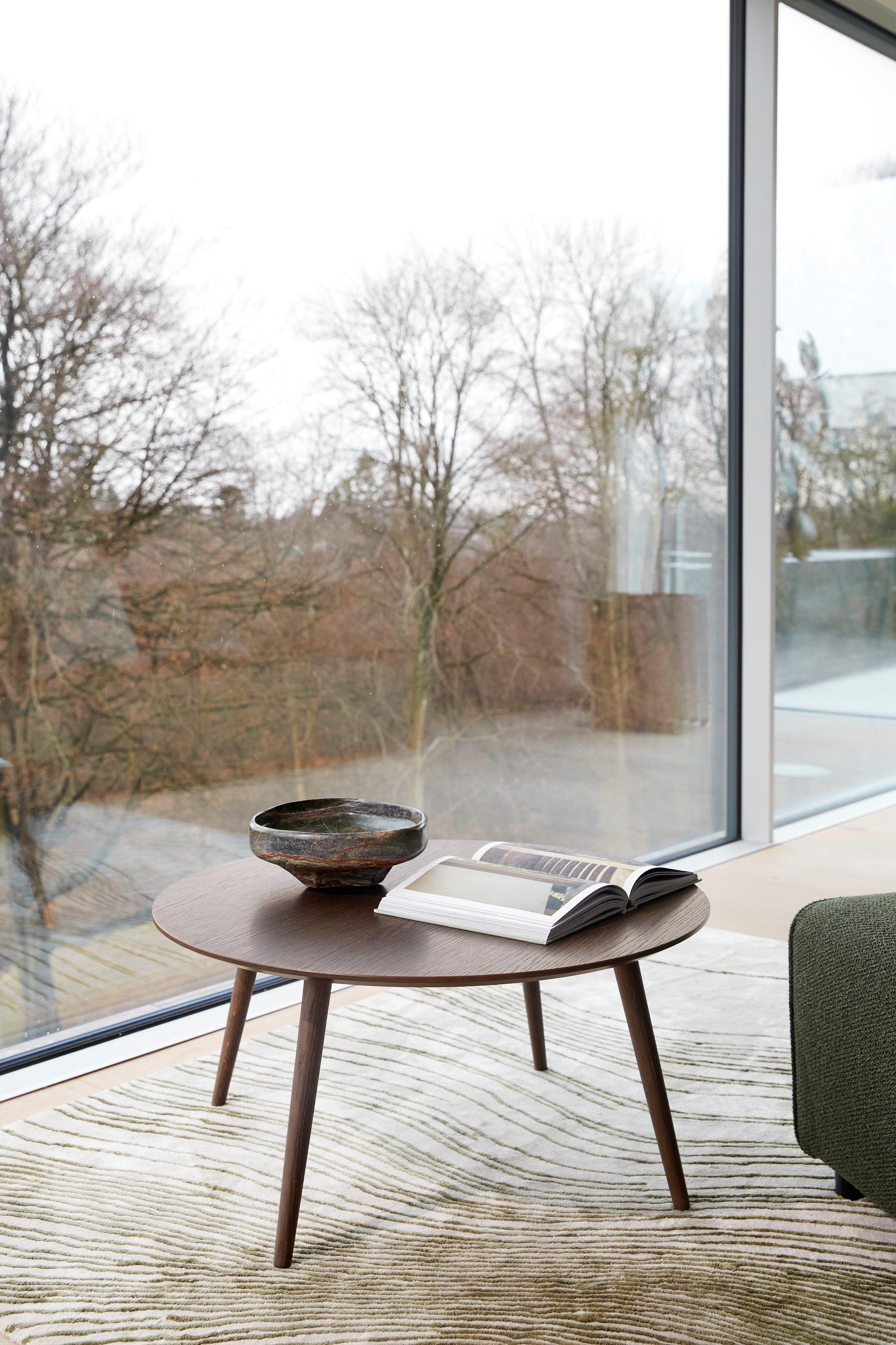 Round Bornholm coffee table with a Curve bowl and open book, placed on a Tide rug in olive colour and a large window overlooking trees.