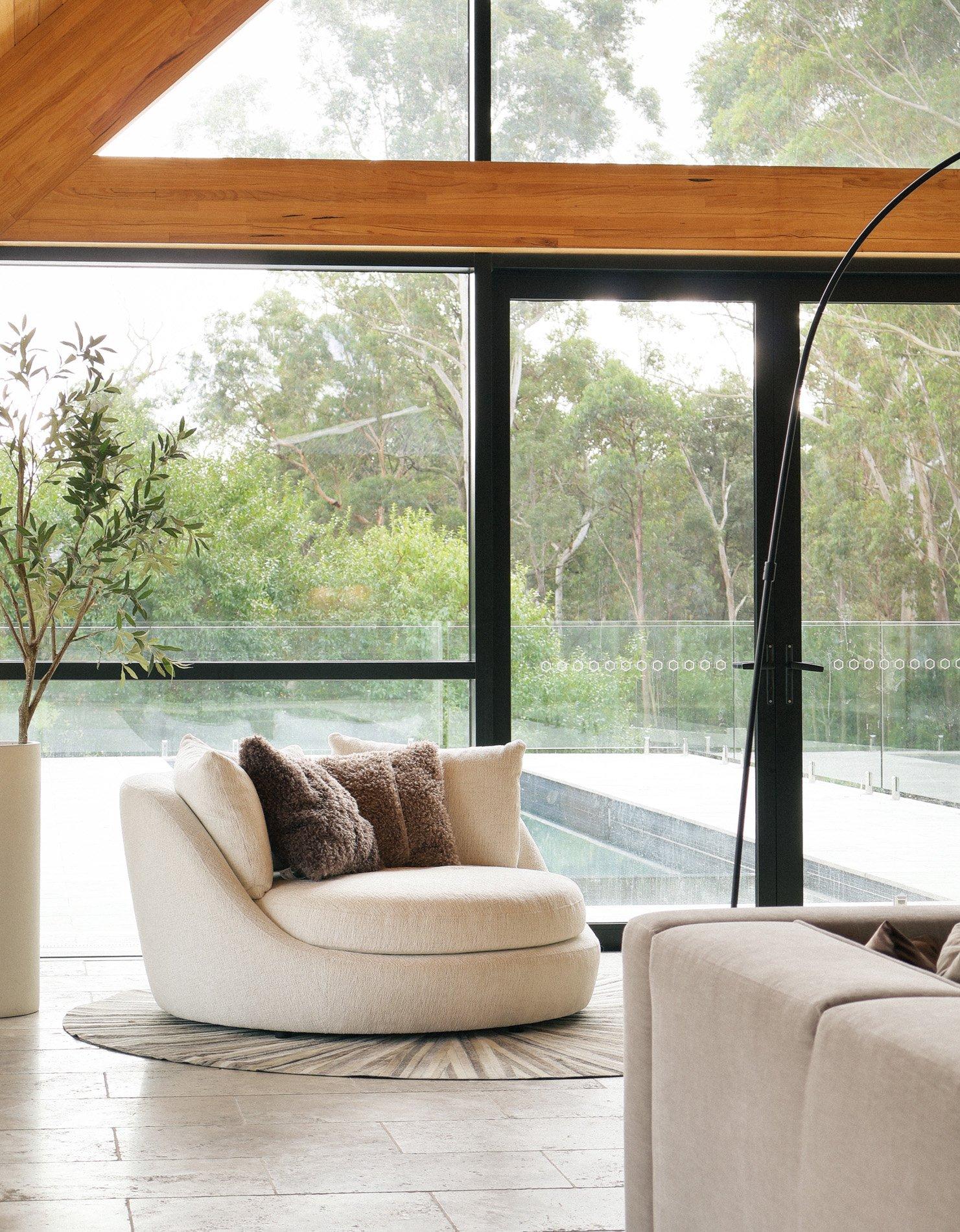 Cream upholstered lounge chair with cushions beside large windows and an arched floor lamp.