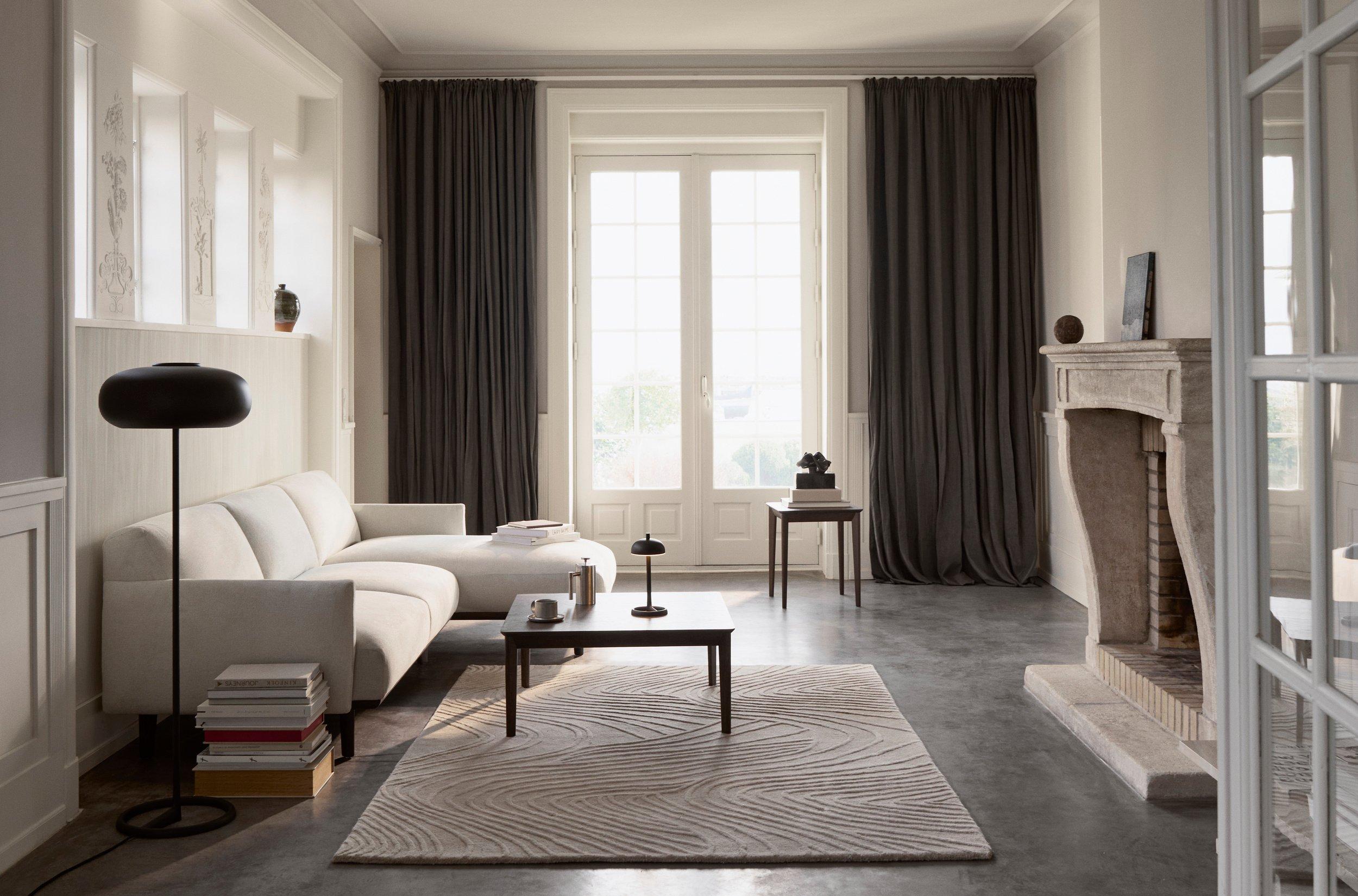 Salon minimaliste avec canapé clair, tables basses et tables d’appoint en chêne foncé, lampes de sol et de table noires et tapis texturé naturel.