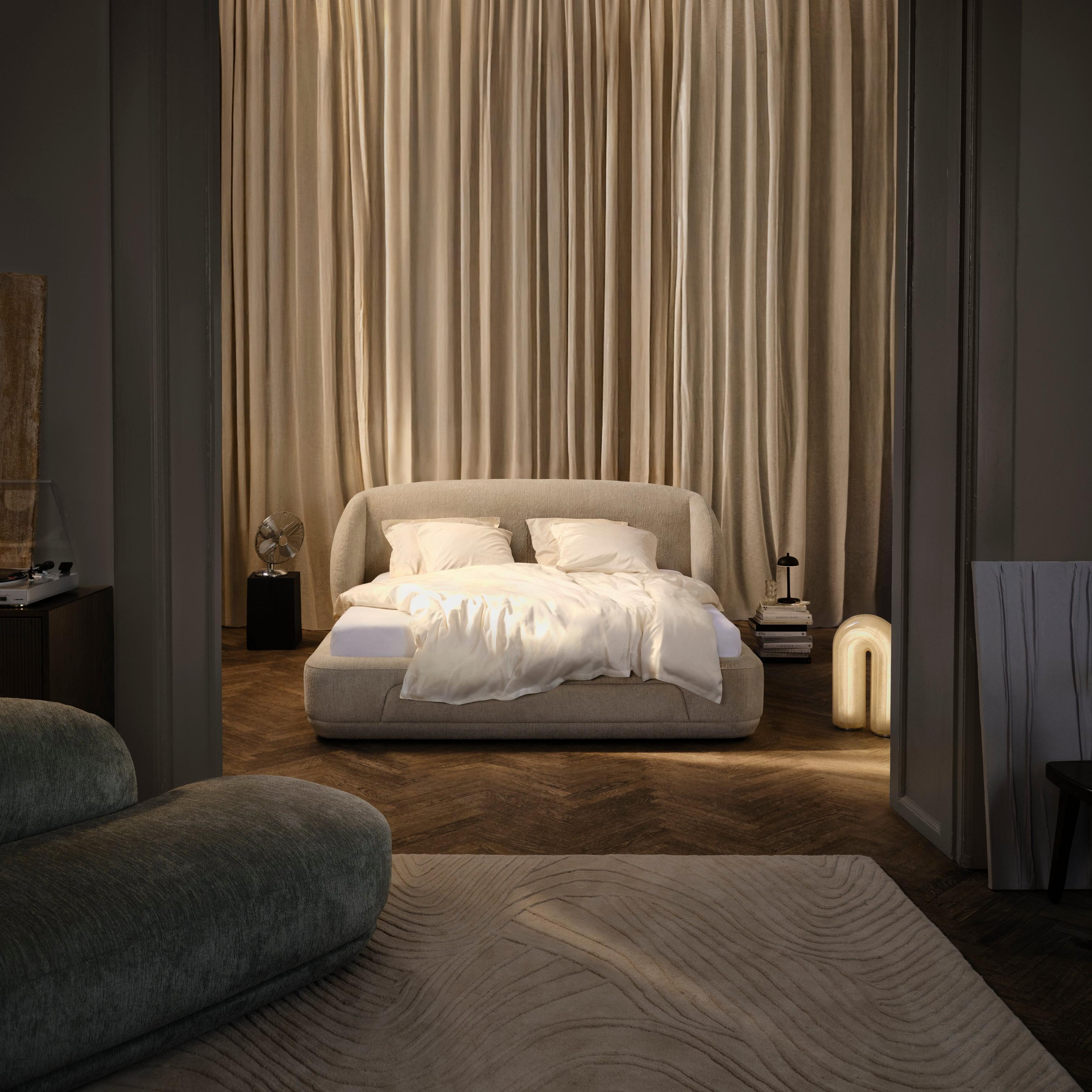 Lit moderne avec draps blancs et coussins, rideaux beiges derrière. Table basse et lampe.