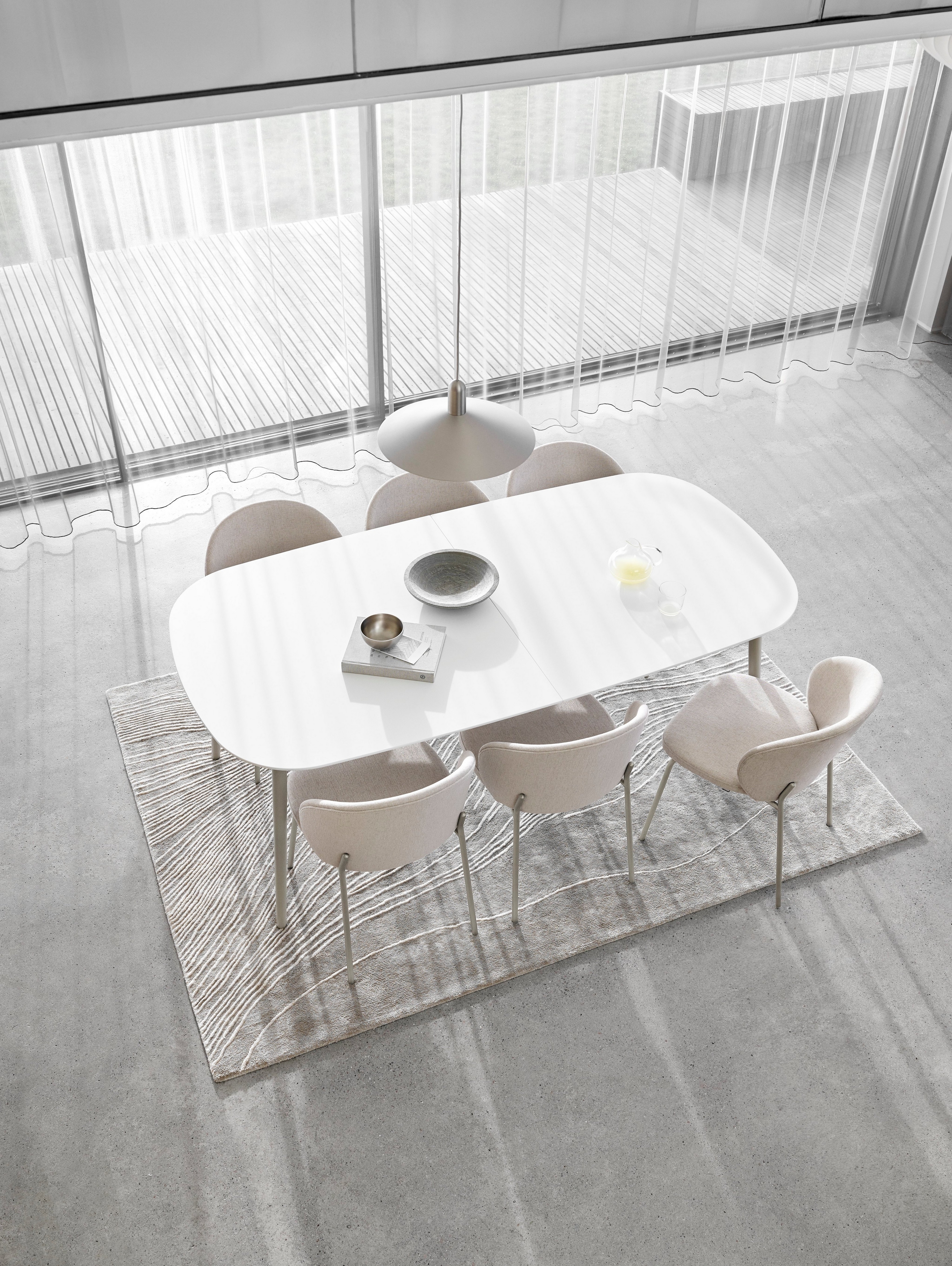 Minimalistyczna jadalnia z białym owalnym stołem, krzesłami, przezroczystymi zasłonami i teksturowanym dywanem.