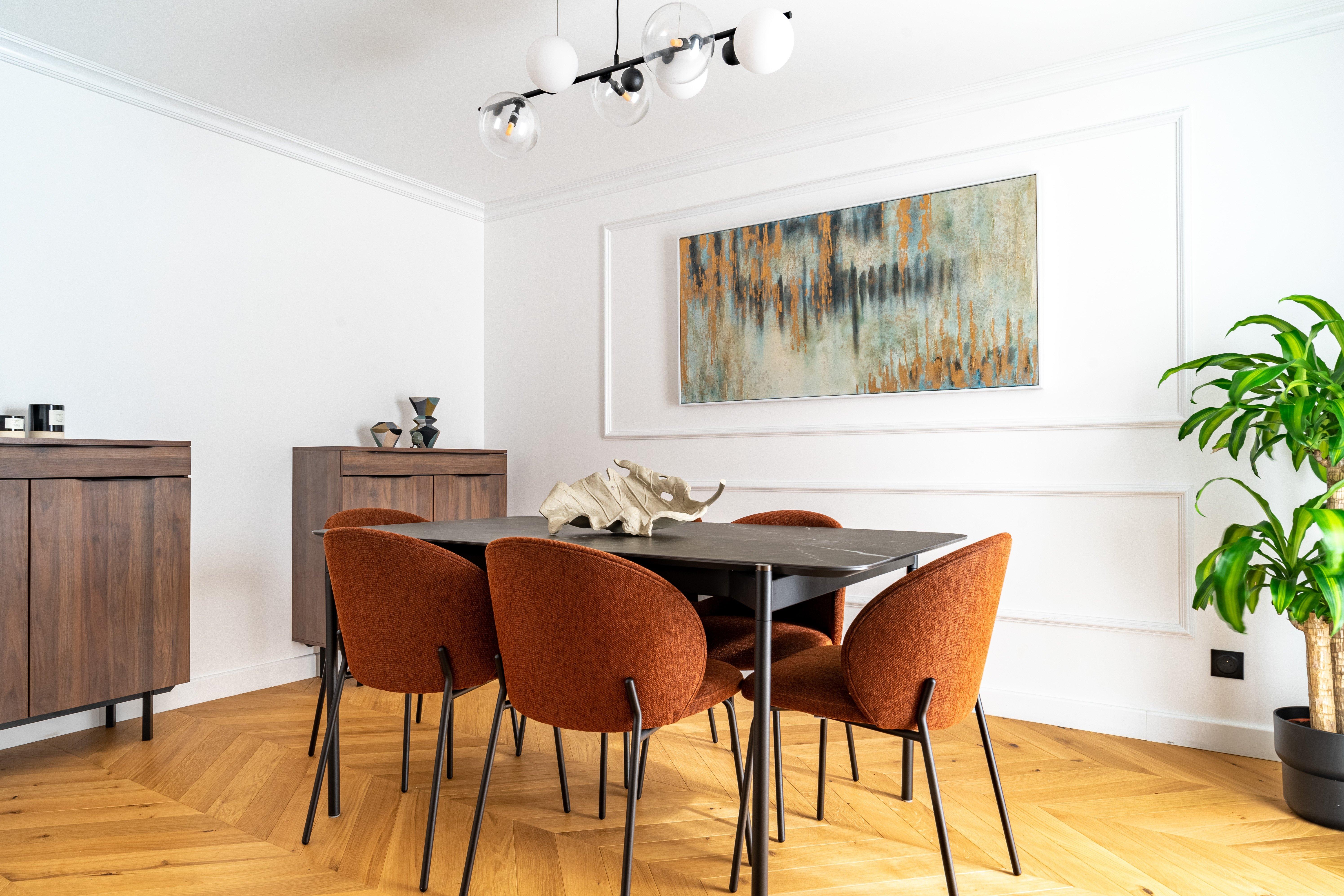Modern dining room with six rust-colored chairs, black table, abstract wall art, wood cabinets, and a large potted plant.