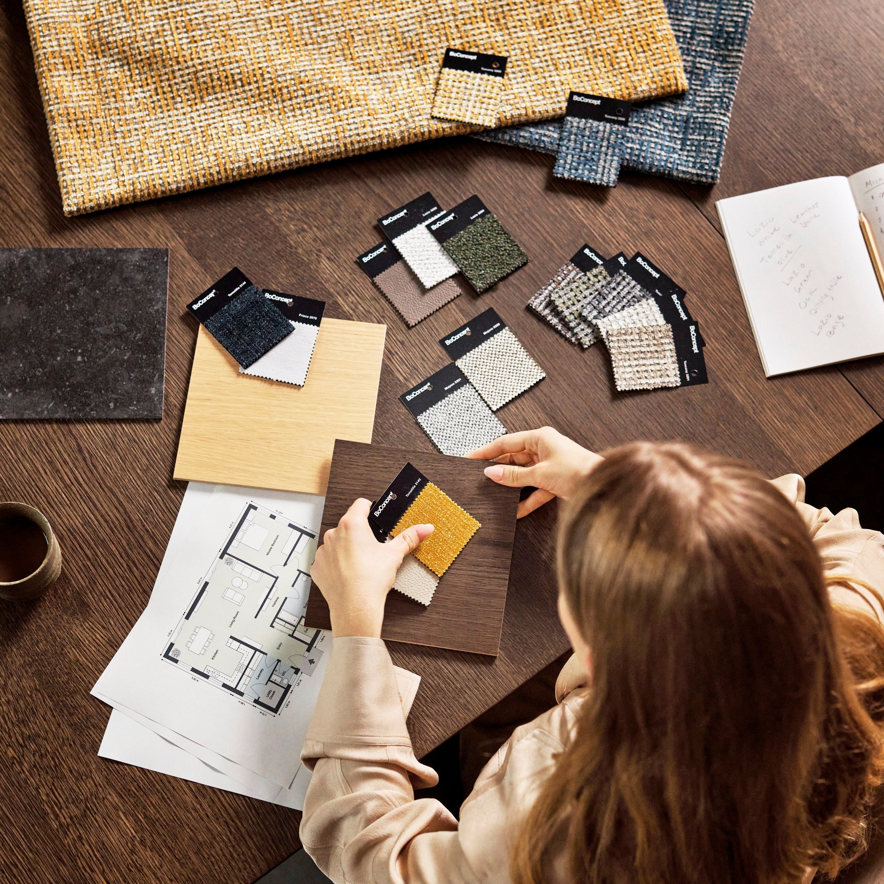 Designer choosing fabric samples on a wooden table with floor plan and notes.
