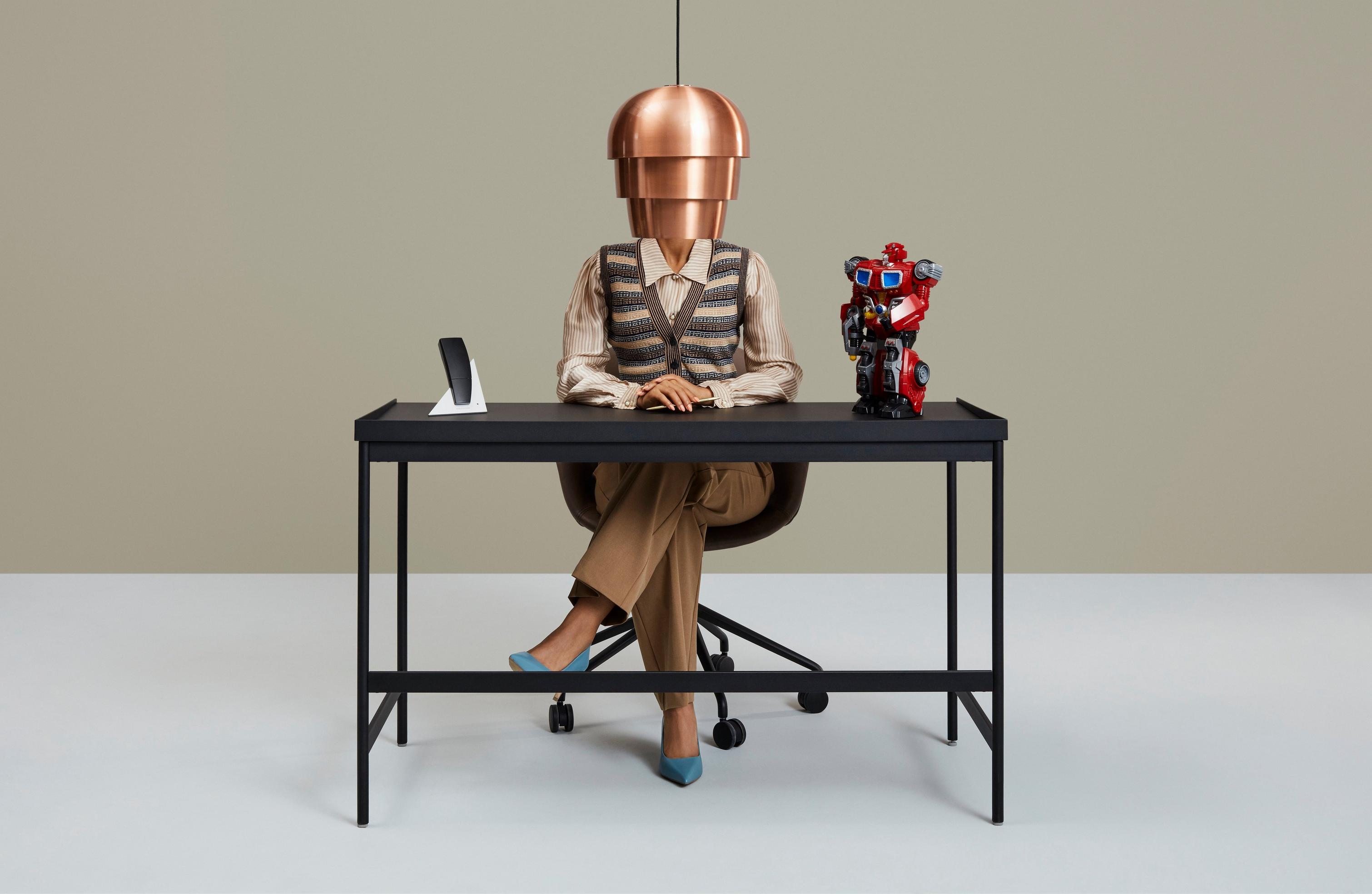 Person at desk with a lampshade headpiece, phone, and colorful robot figurine.