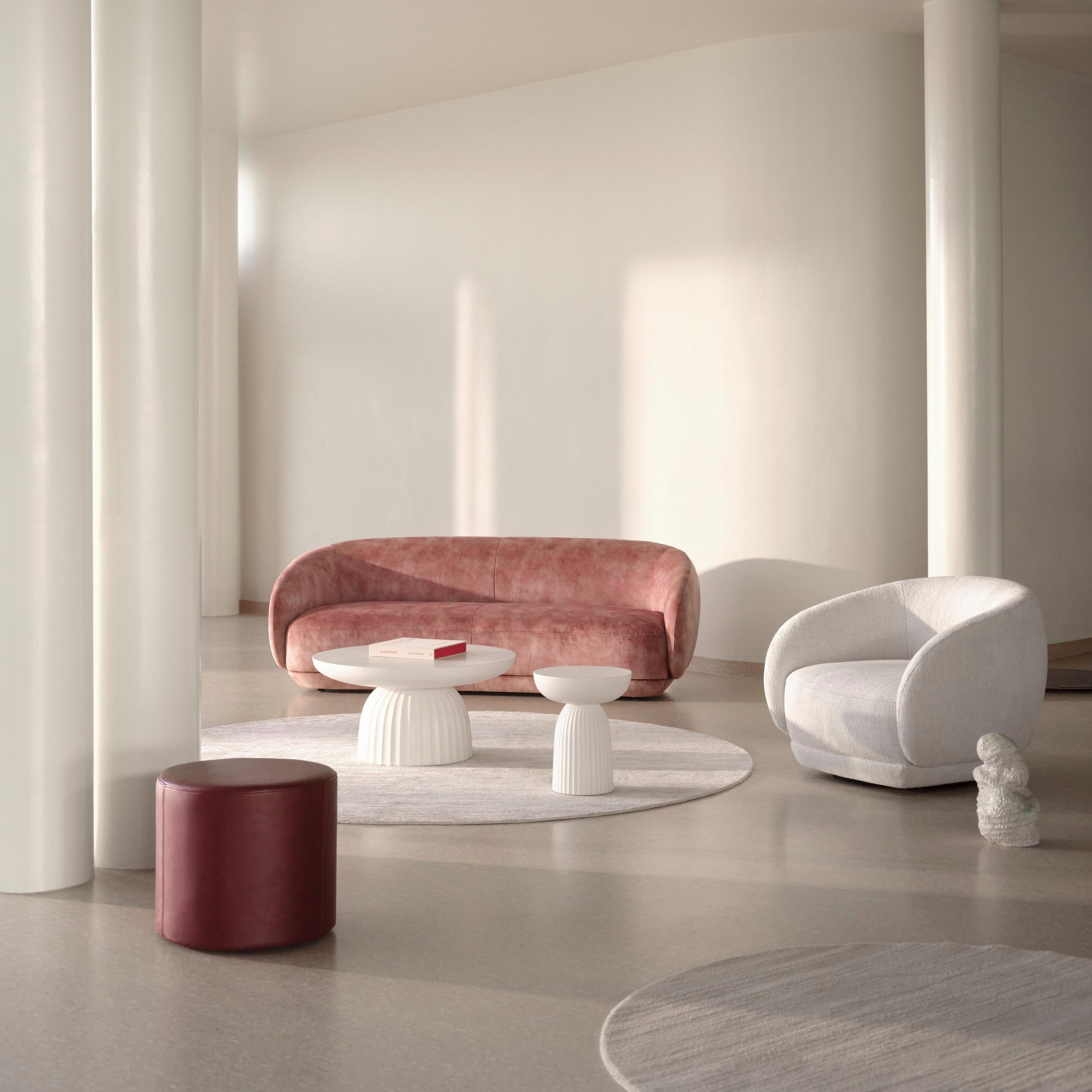 Minimalist lounge with blush pink sofa, white curved armchair, fluted white coffee tables, round rugs, and burgundy cylindrical pouf.