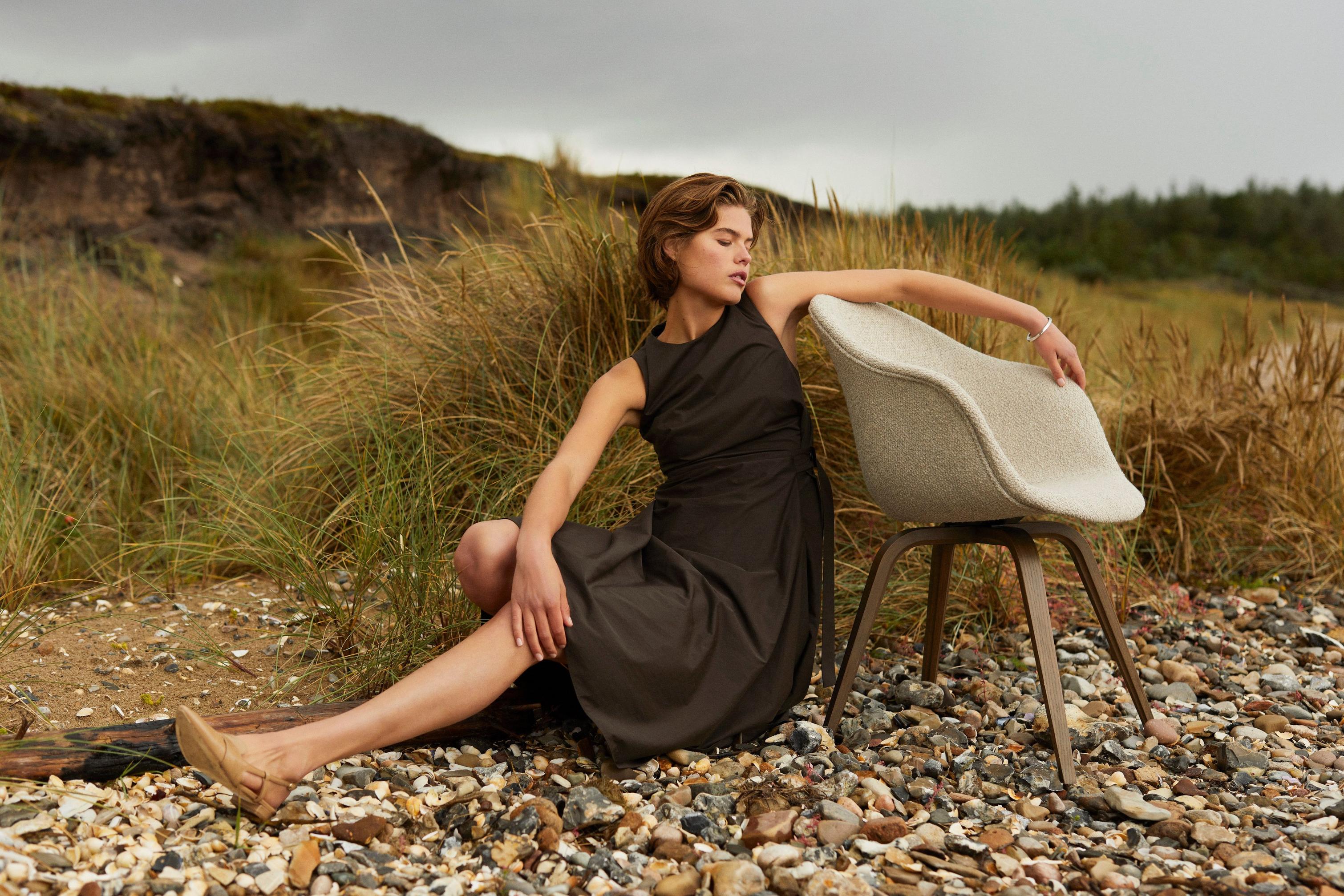 Nainen nojaa beigellä Lazio-kankaalla verhoiltuun Hauge-ruokapöydän tuoliin niityllä rannan lähellä.