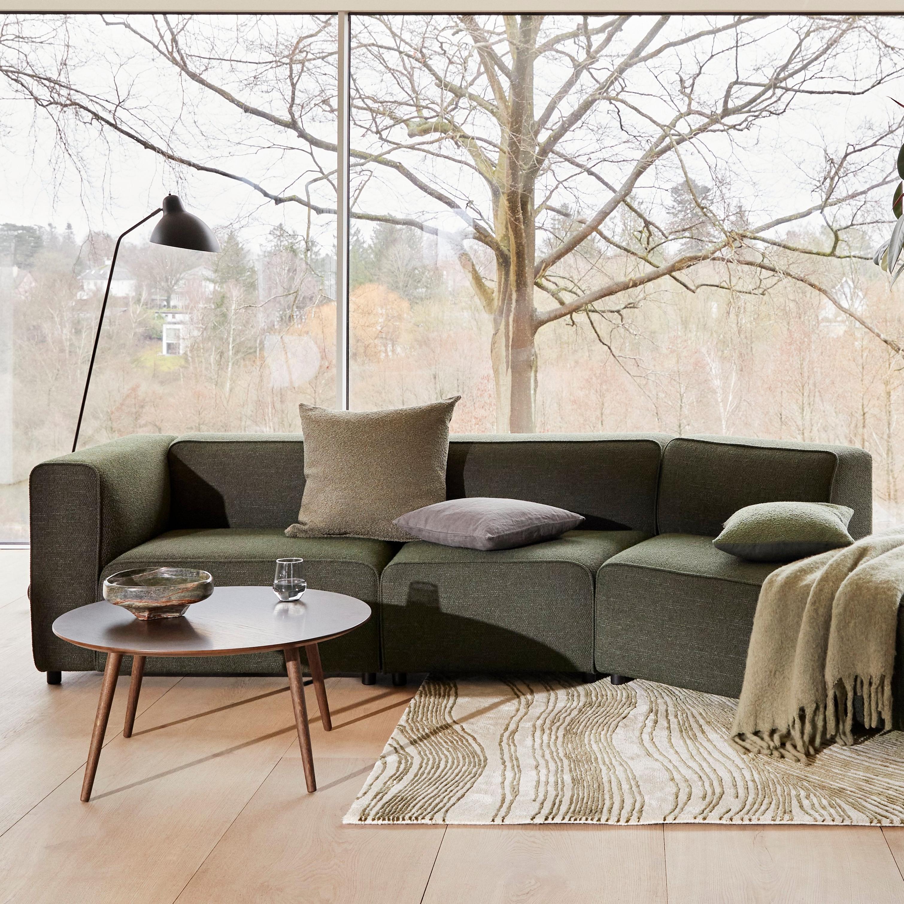 Living room with a green Carmo sofa, assorted pillows, round Bornholm coffee table, Demand floor lamp, potted plant, and large windows with a tree view.
