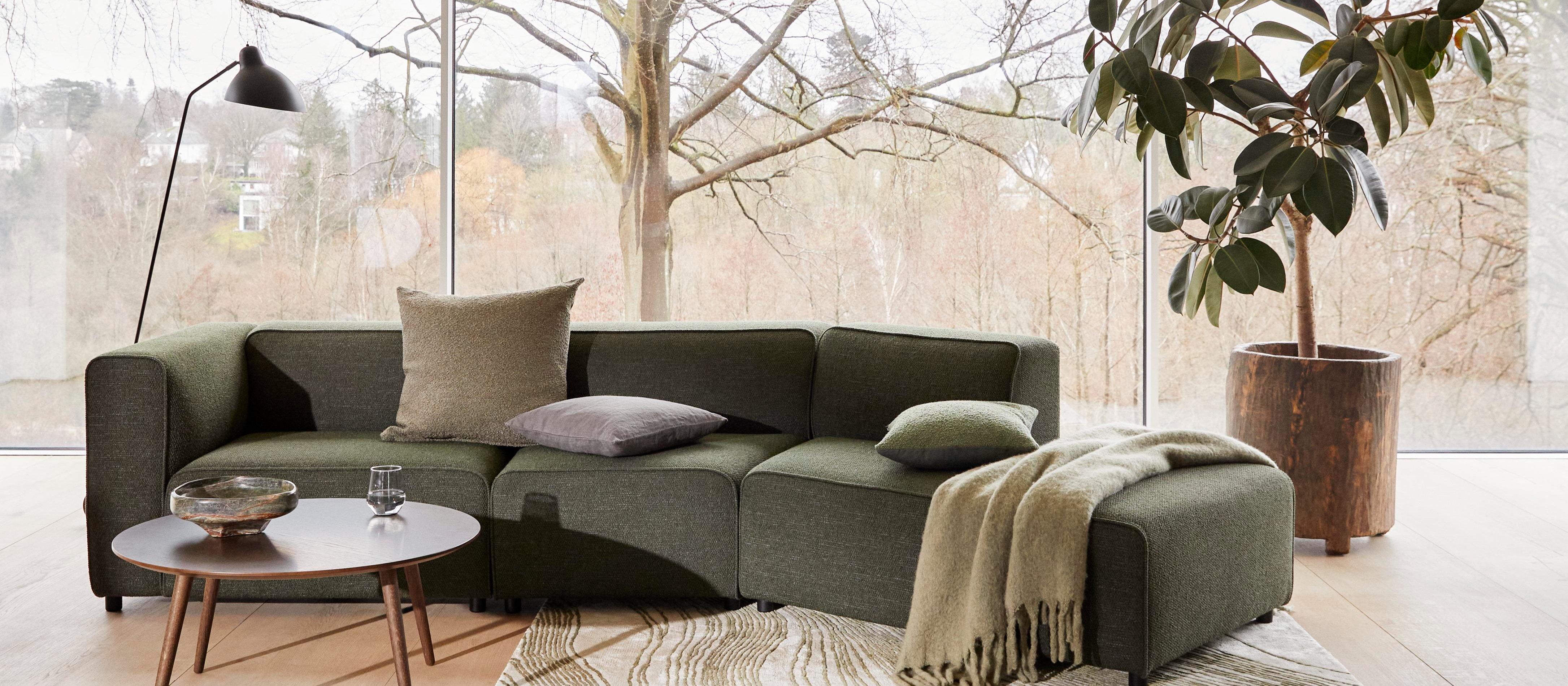 Salon avec un canapé Carmo vert, des coussins assortis, une table basse ronde Bornholm, une lampe de sol Demand, une plante en pot et de grandes fenêtres avec vue sur l’arbre.
