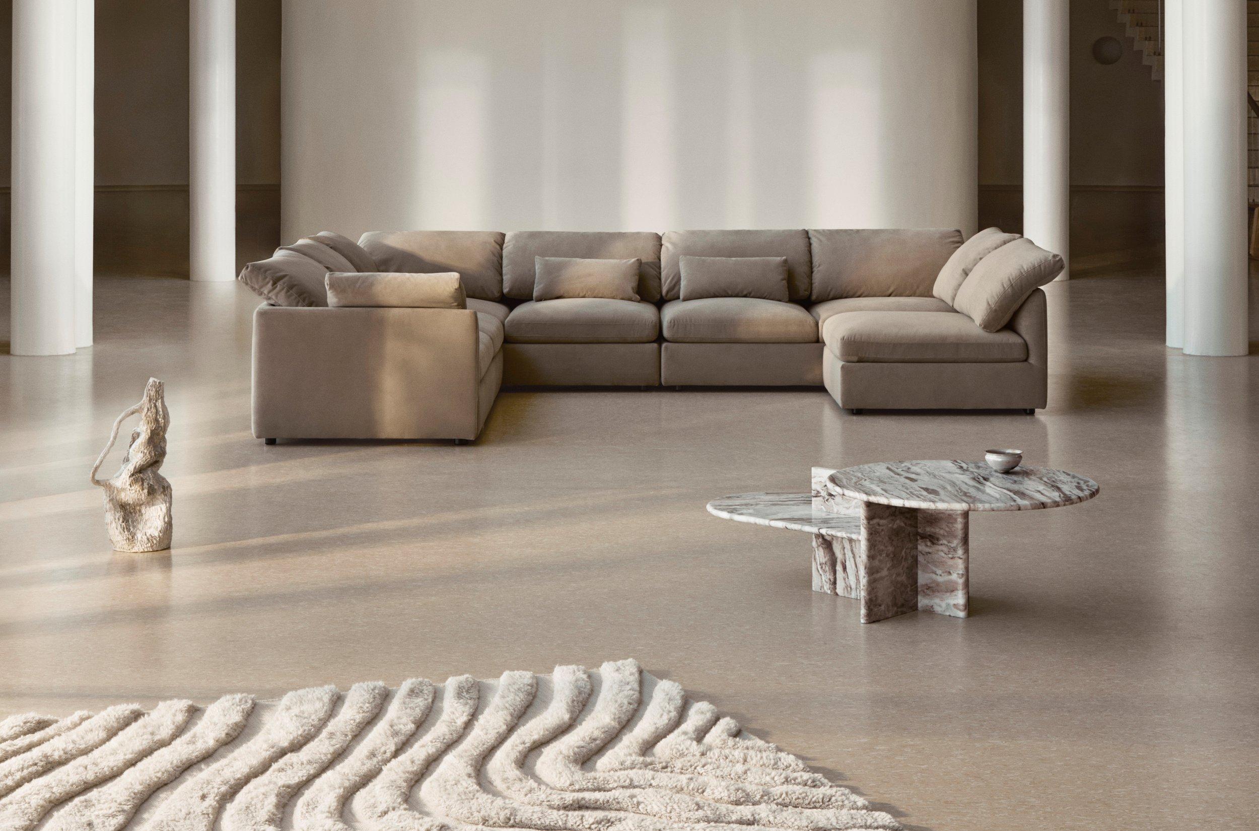 Espacio amplio con sofá modular en tela arena, mesa de centro redonda de mármol marrón y alfombras texturizadas en crema y gris.