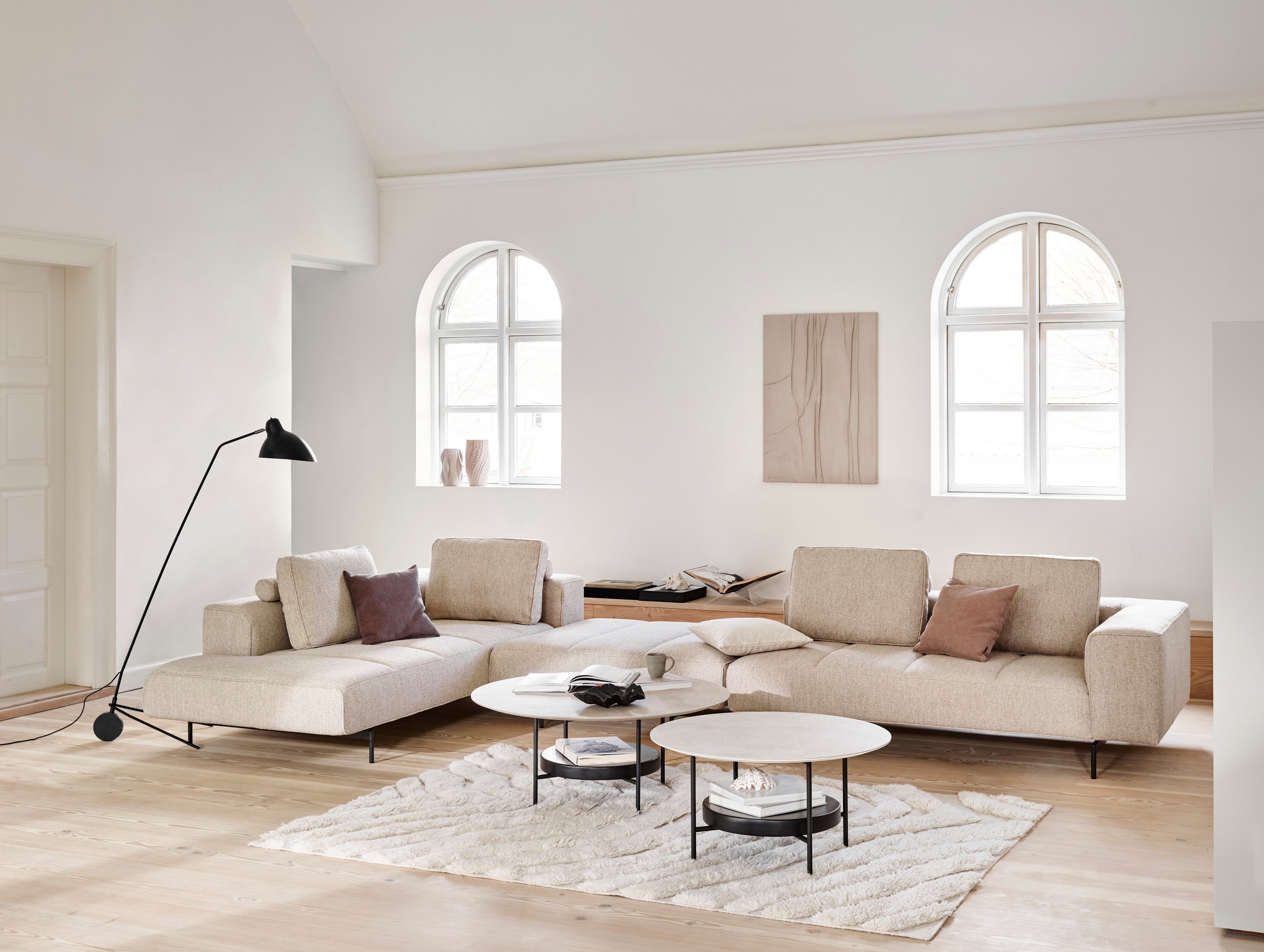 Sala de estar minimalista com sofá modular Amsterdam, mesas de centro Madrid, candeeiro de chão e janelas arqueadas.