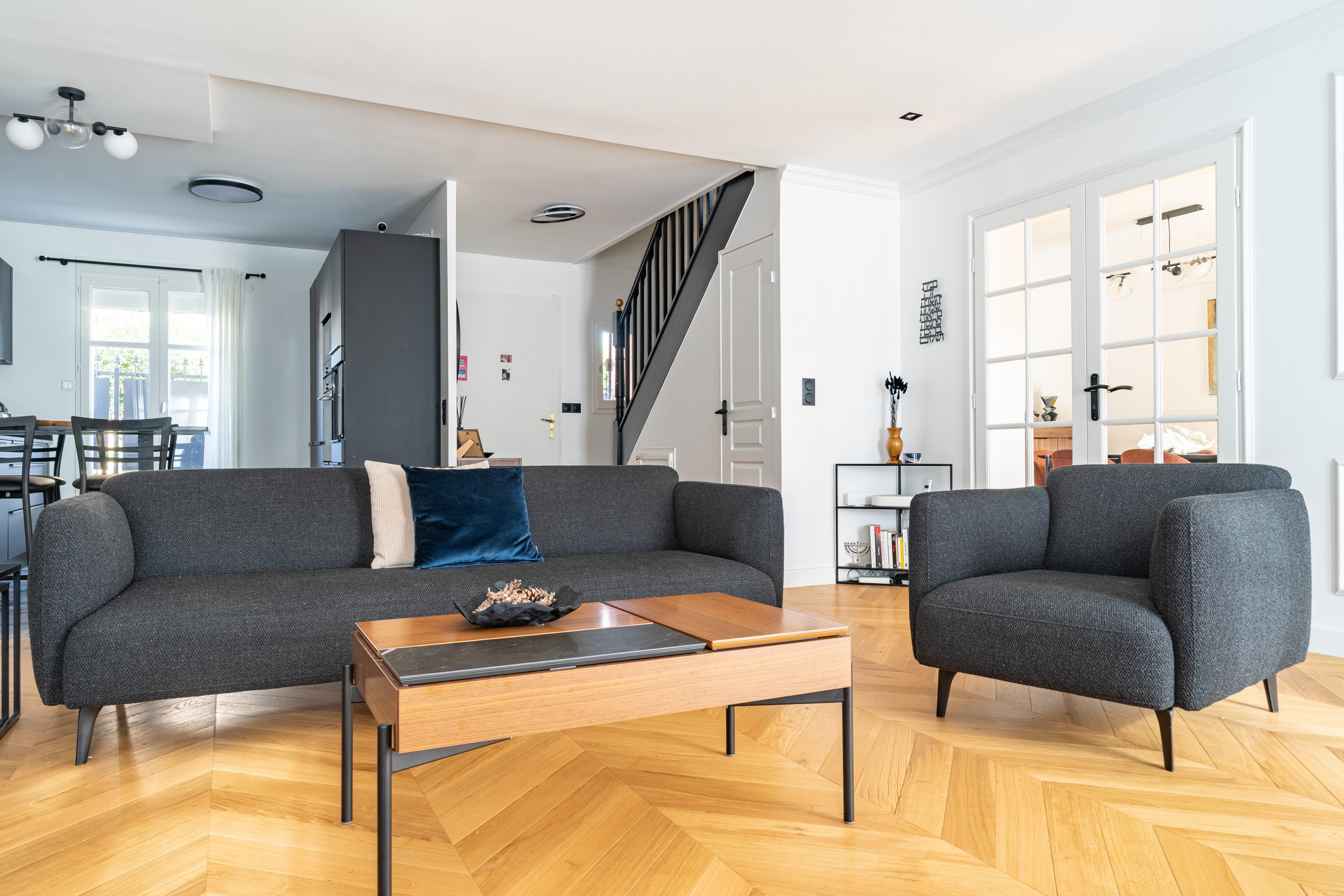 Open-plan space with dark grey sofa set, wood coffee table, chevron flooring, and view into kitchen and dining areas.