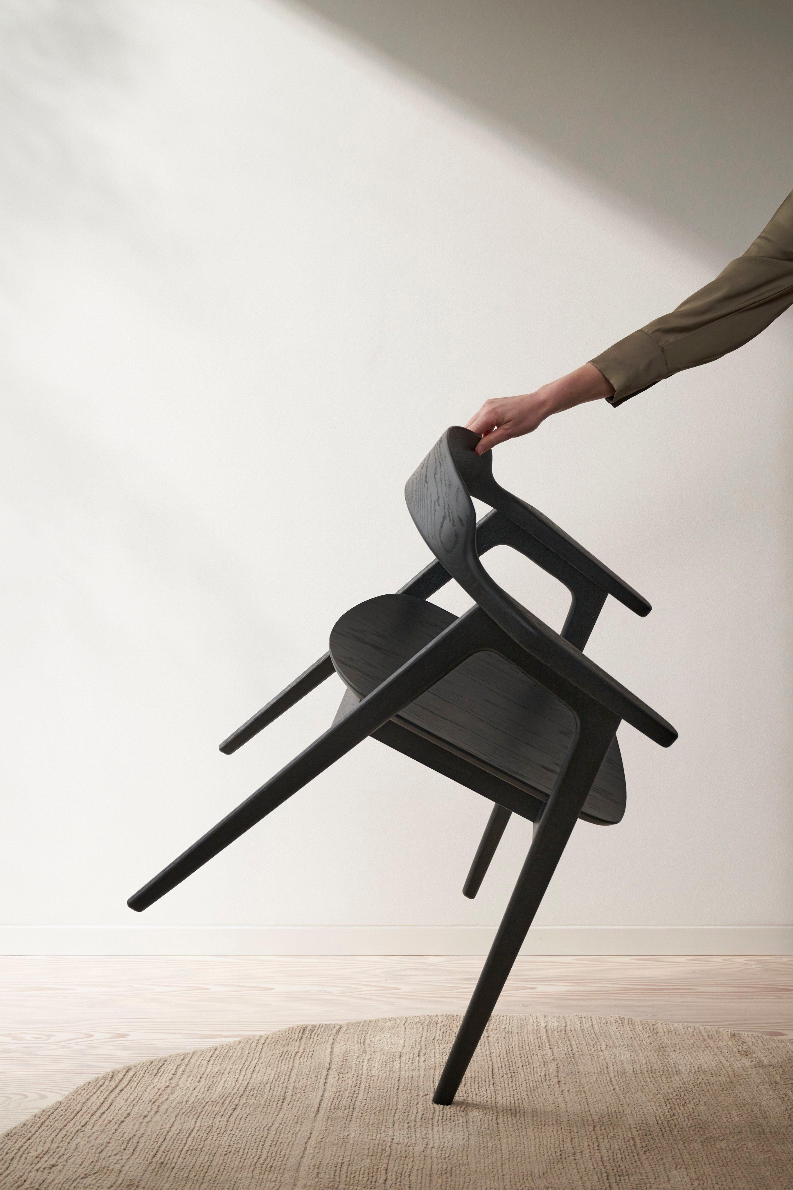 Person, die einen schwarzen, minimalistischen Seoul Esszimmerstuhl aus Holz diagonal geneigt an der Rückenlehne hält. Das Sonnenlicht wirft Schatten.