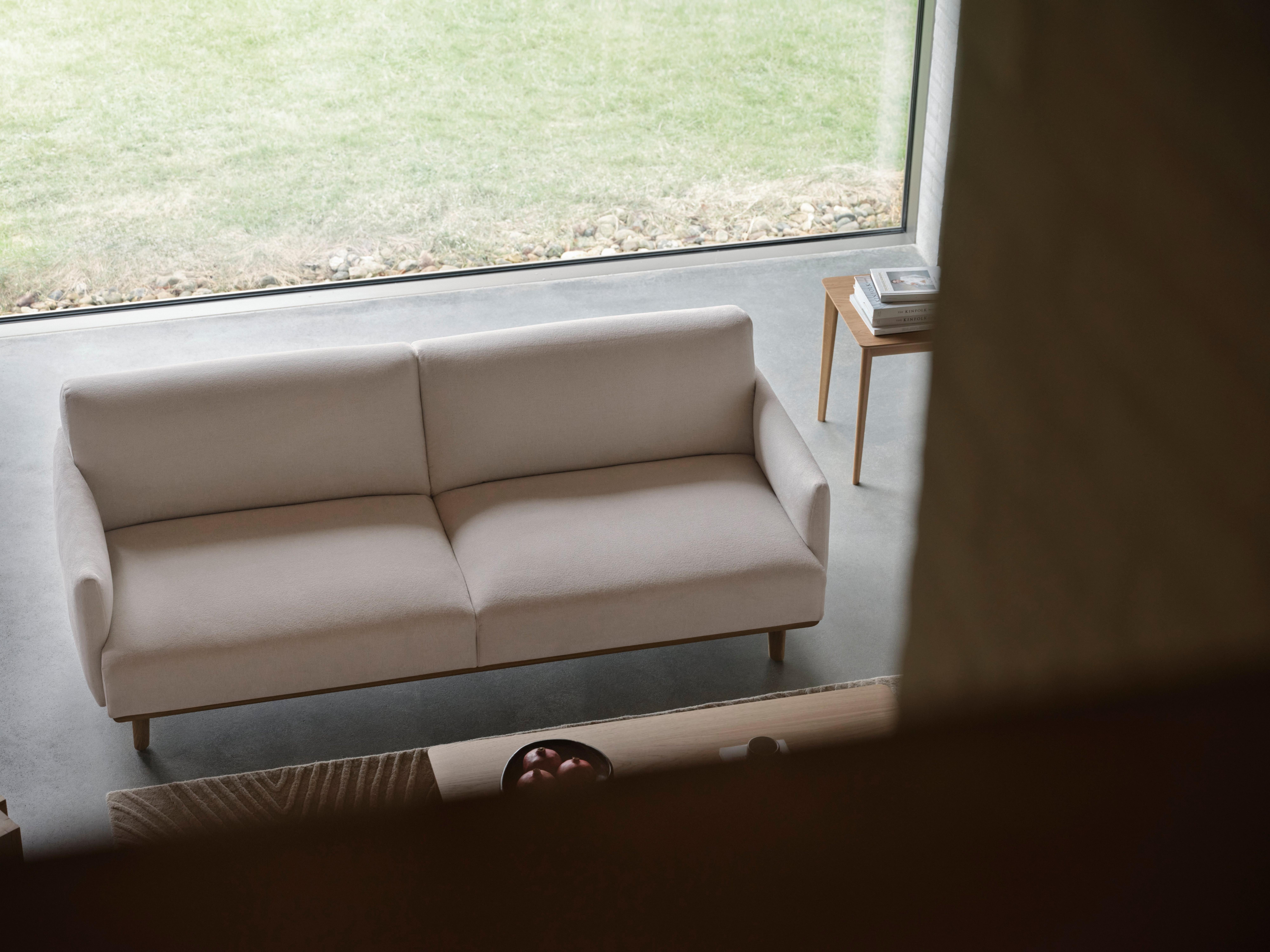 Bílá dvoumístná pohovka s dřevěnými nohami umístěná vedle světlého dřevěného odkládacího stolku s knihami nedaleko velkého okna.