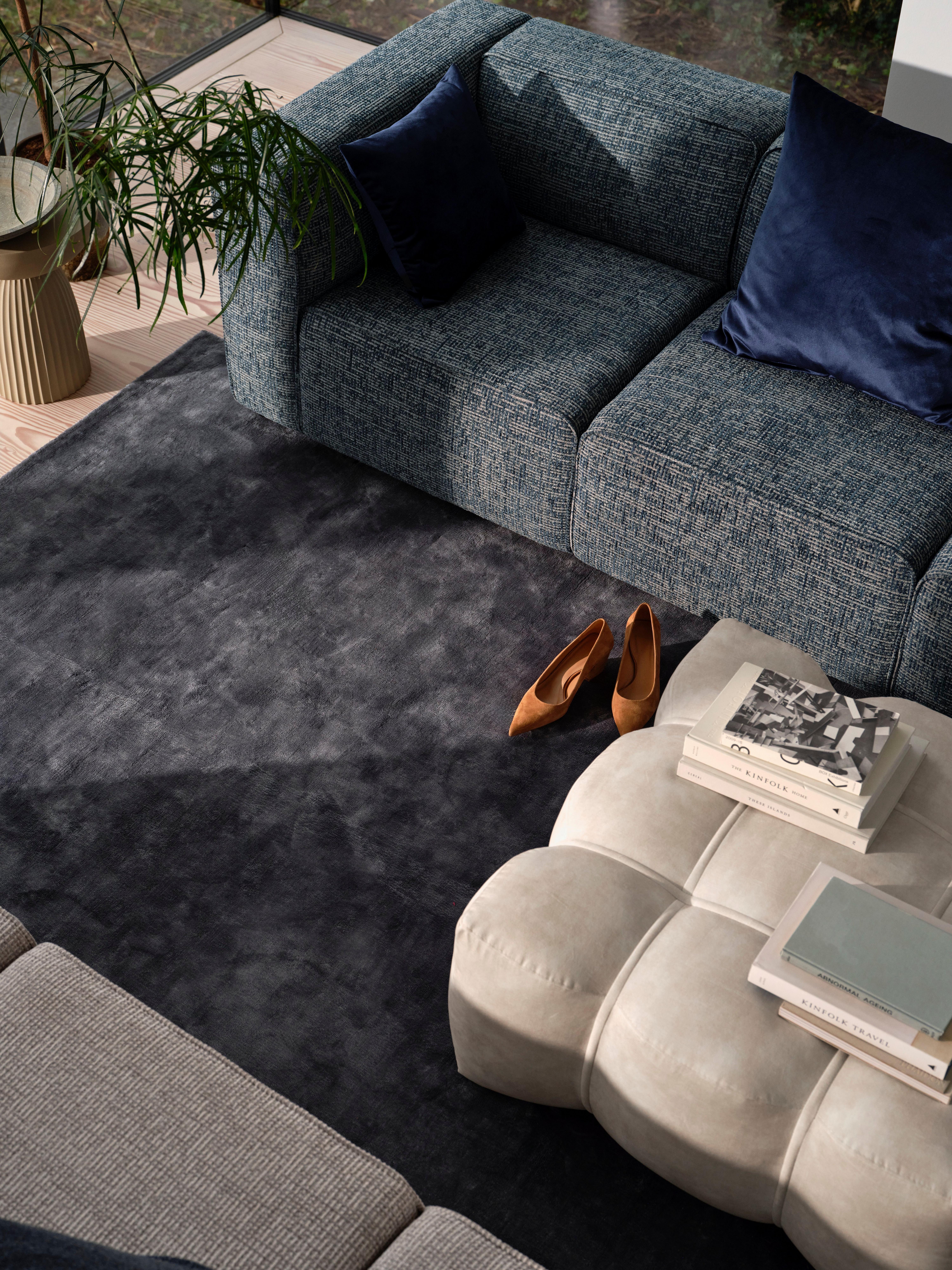 Salon avec canapé bleu, coussins marine, repose-pieds Nawabari beige, livres, plante en pot et chaussures marron sur tapis gris foncé.