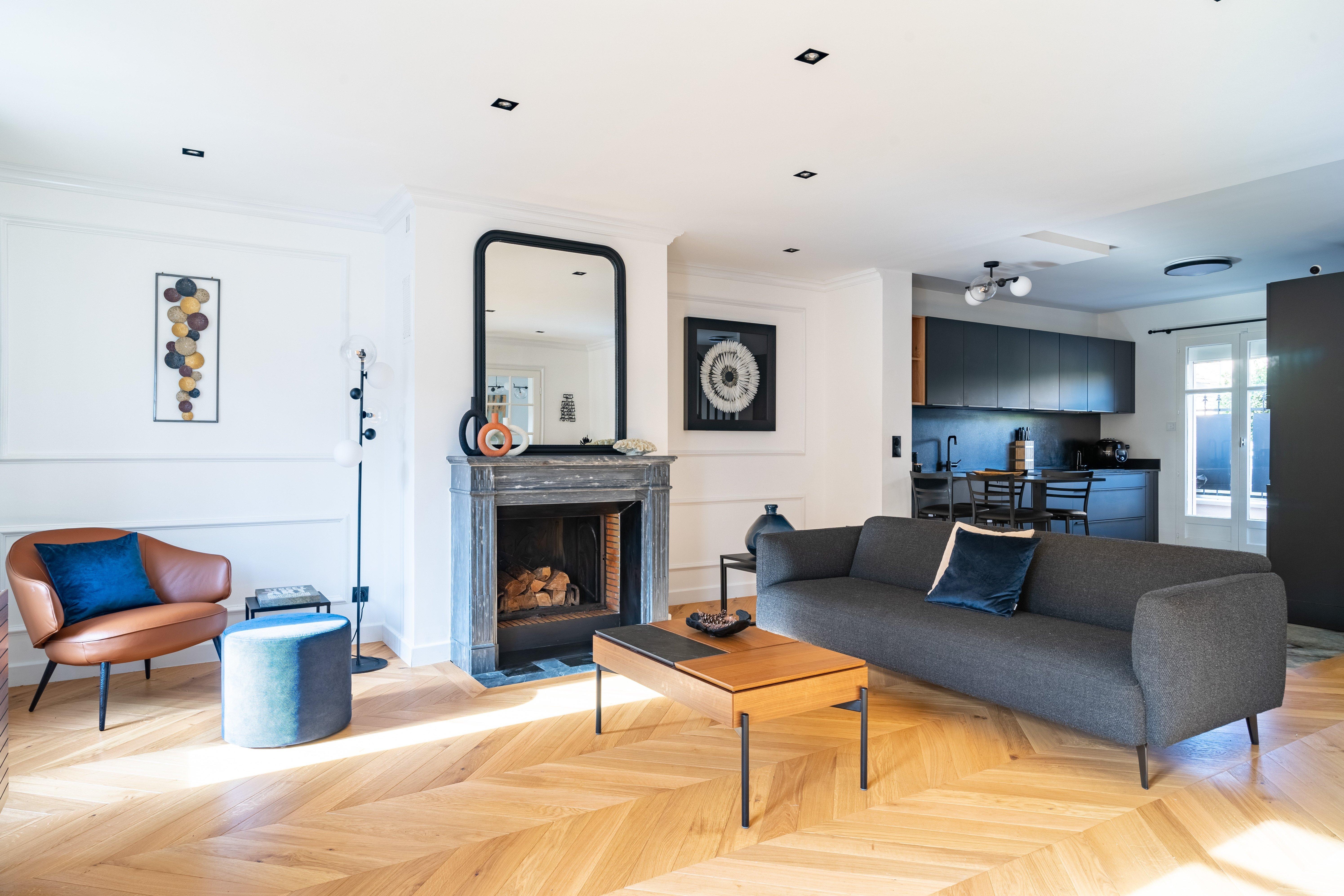 Modern living room with grey sofa, brown chair, fireplace, and wooden coffee table. Kitchen in background.