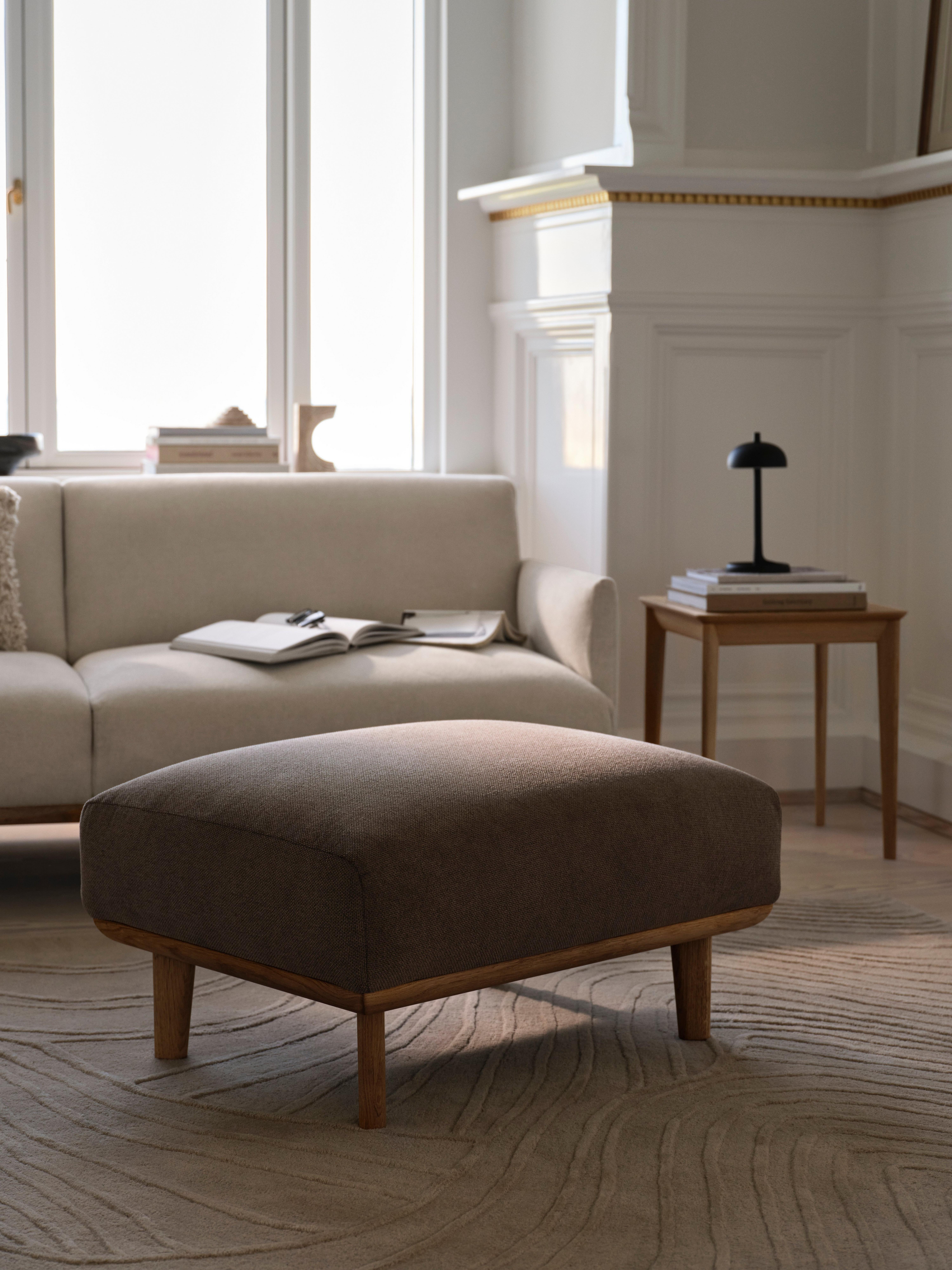Grey ottoman on patterned rug, near a beige sofa with an open book and a lamp on a side table.