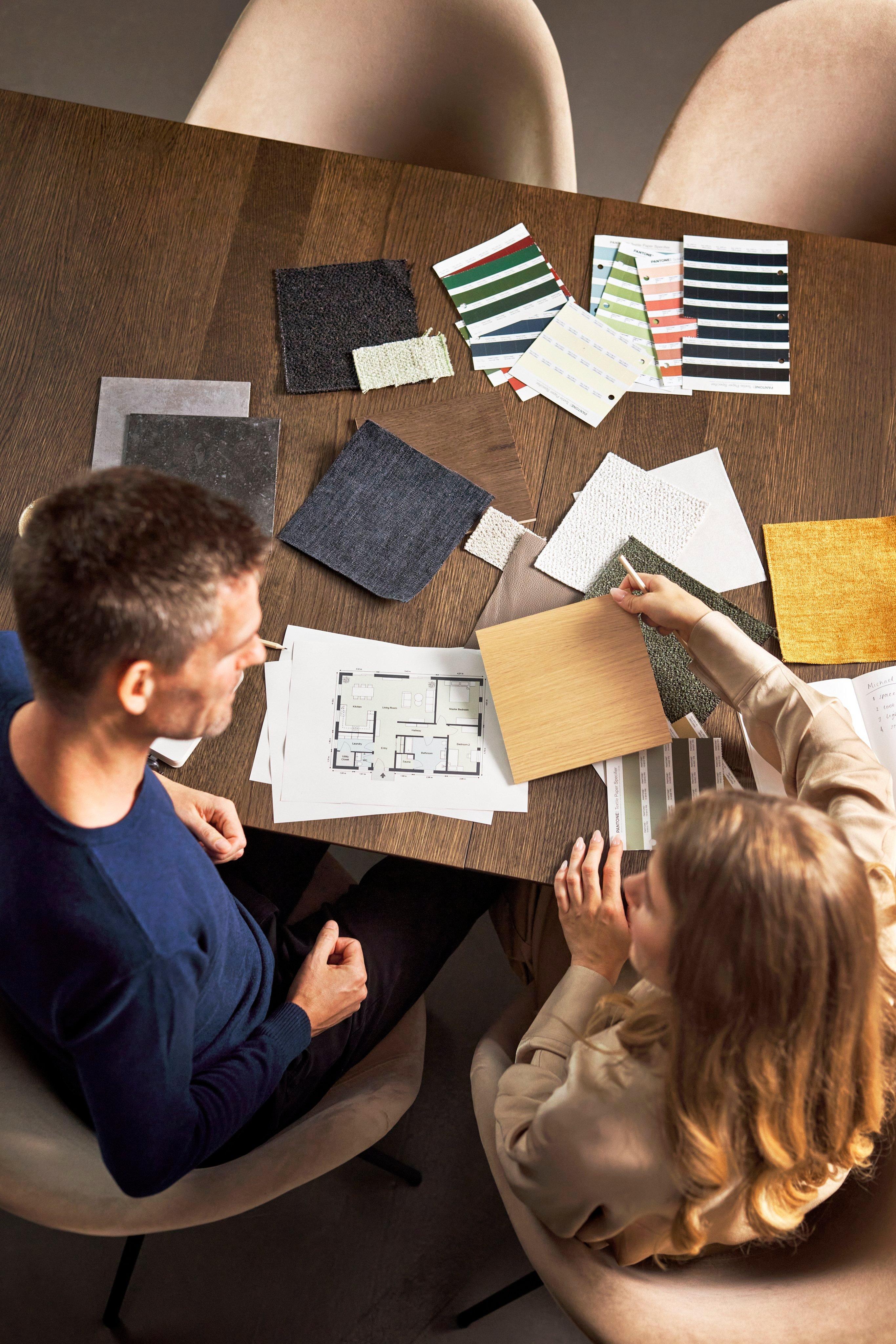 Pohled shora na dvě osoby diskutující o vzorcích materiálů nad stolem s půdorysy.