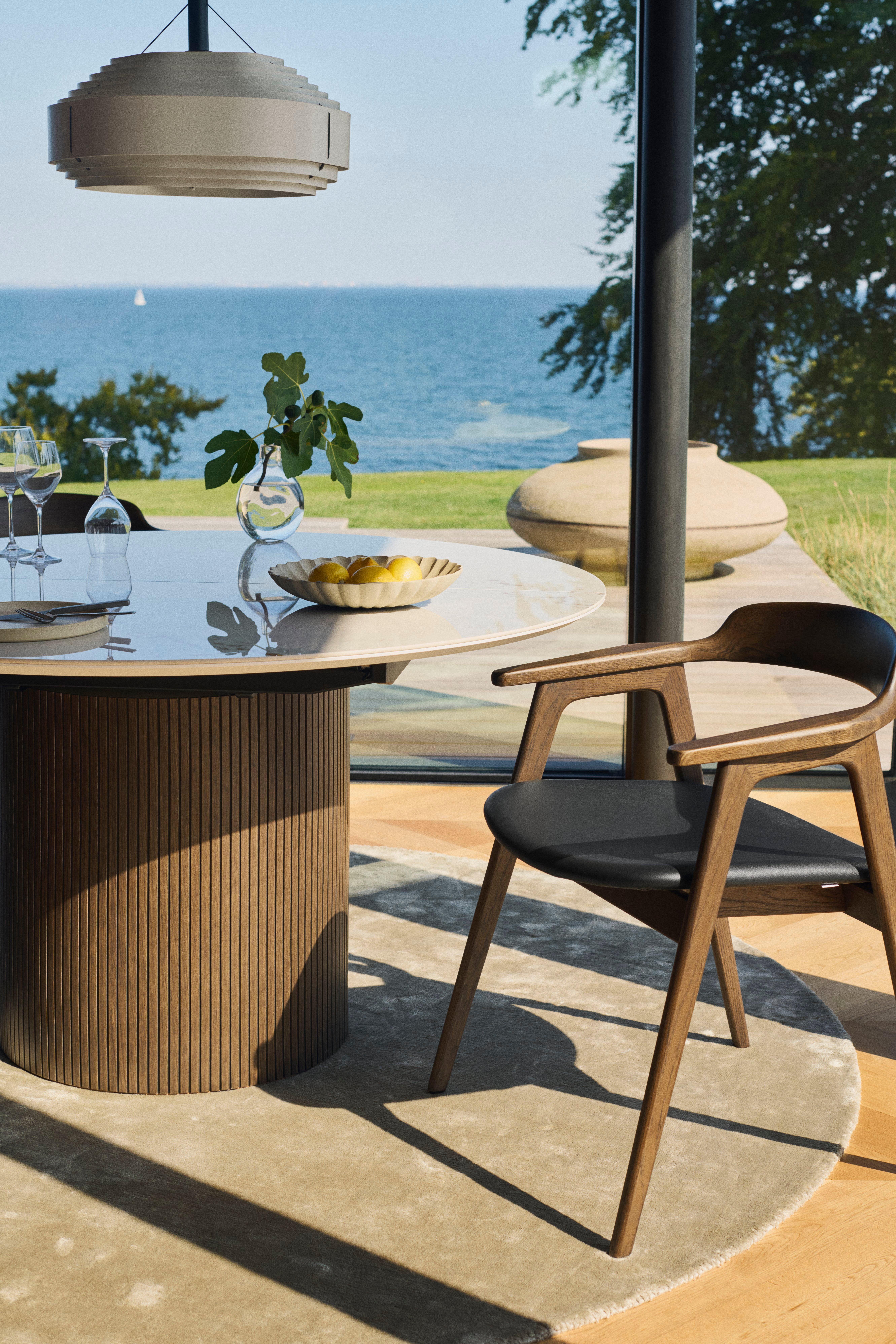 Modern dining area with round wooden table, four chairs, and pendant light. Floor-to-ceiling windows showcase a seaside view.