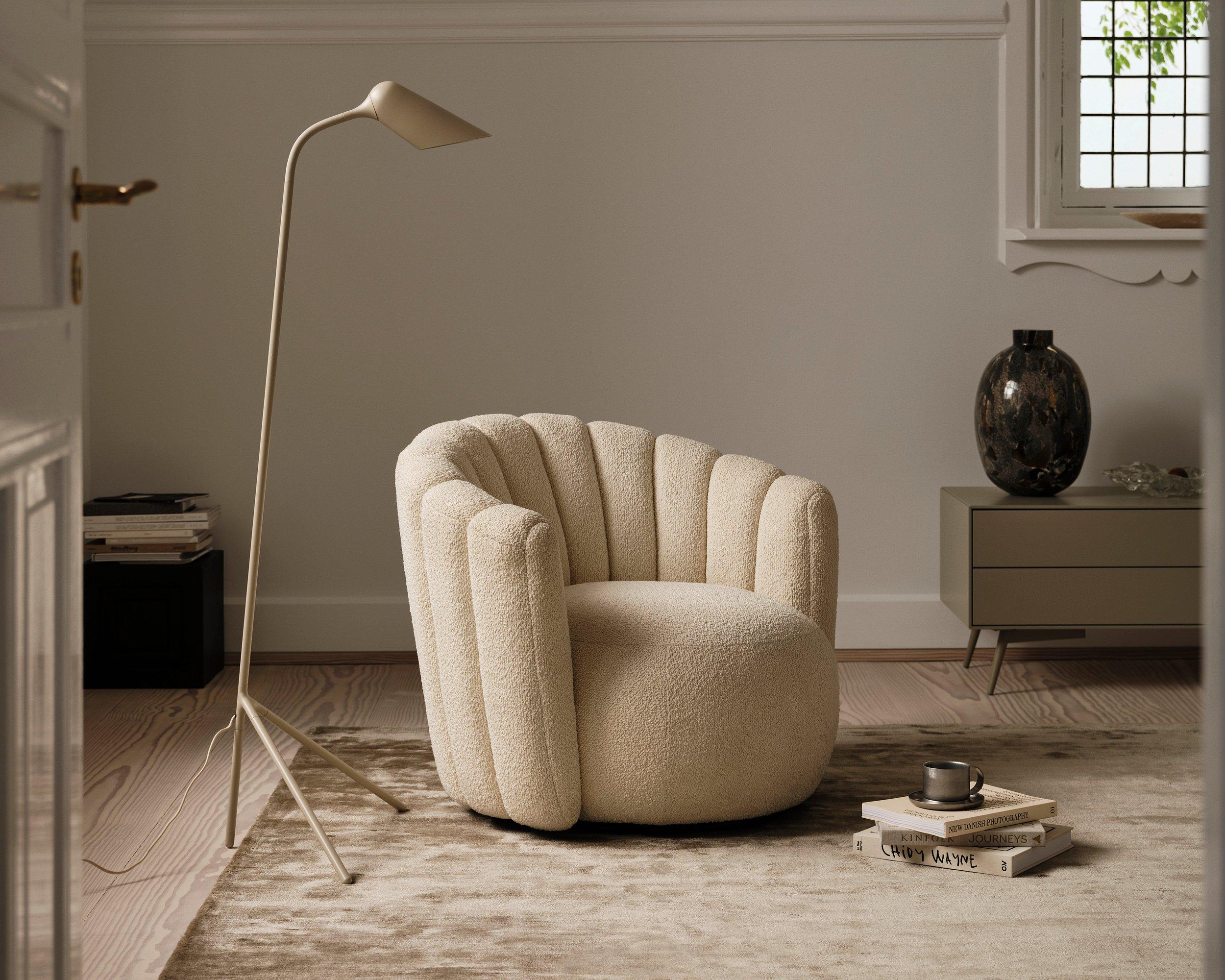 Beige armchair with a scalloped design, floor lamp, books, and a vase on a side table.