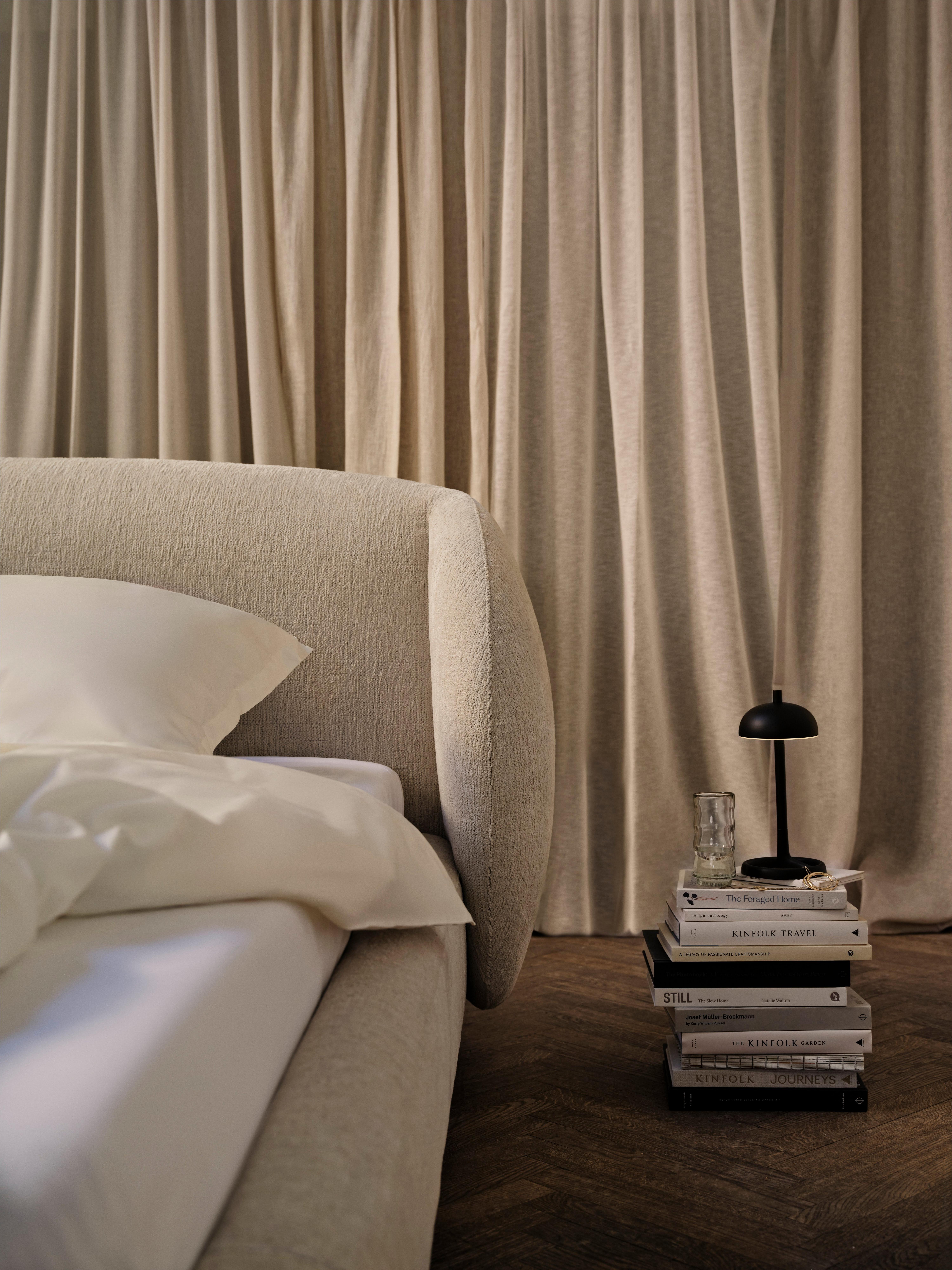 Bolzano bed in beige Lucca fabric with slats, next to stacked books and a matt black Paris table lamp.