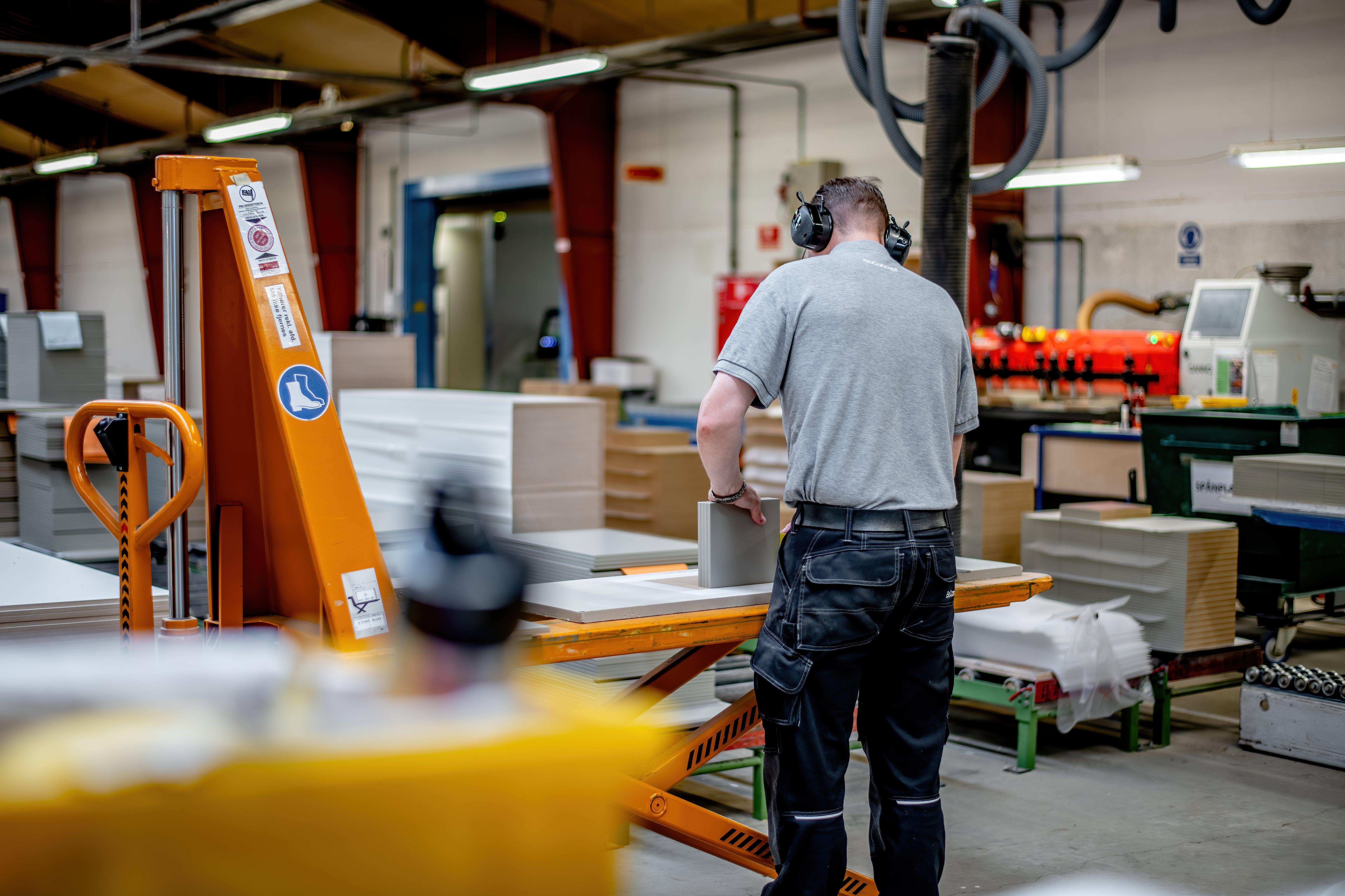 Un trabajador con auriculares maneja maquinaria en una fábrica, manipulando paneles de madera. Cerca se encuentra un elevador de estibas y pilas de materiales.