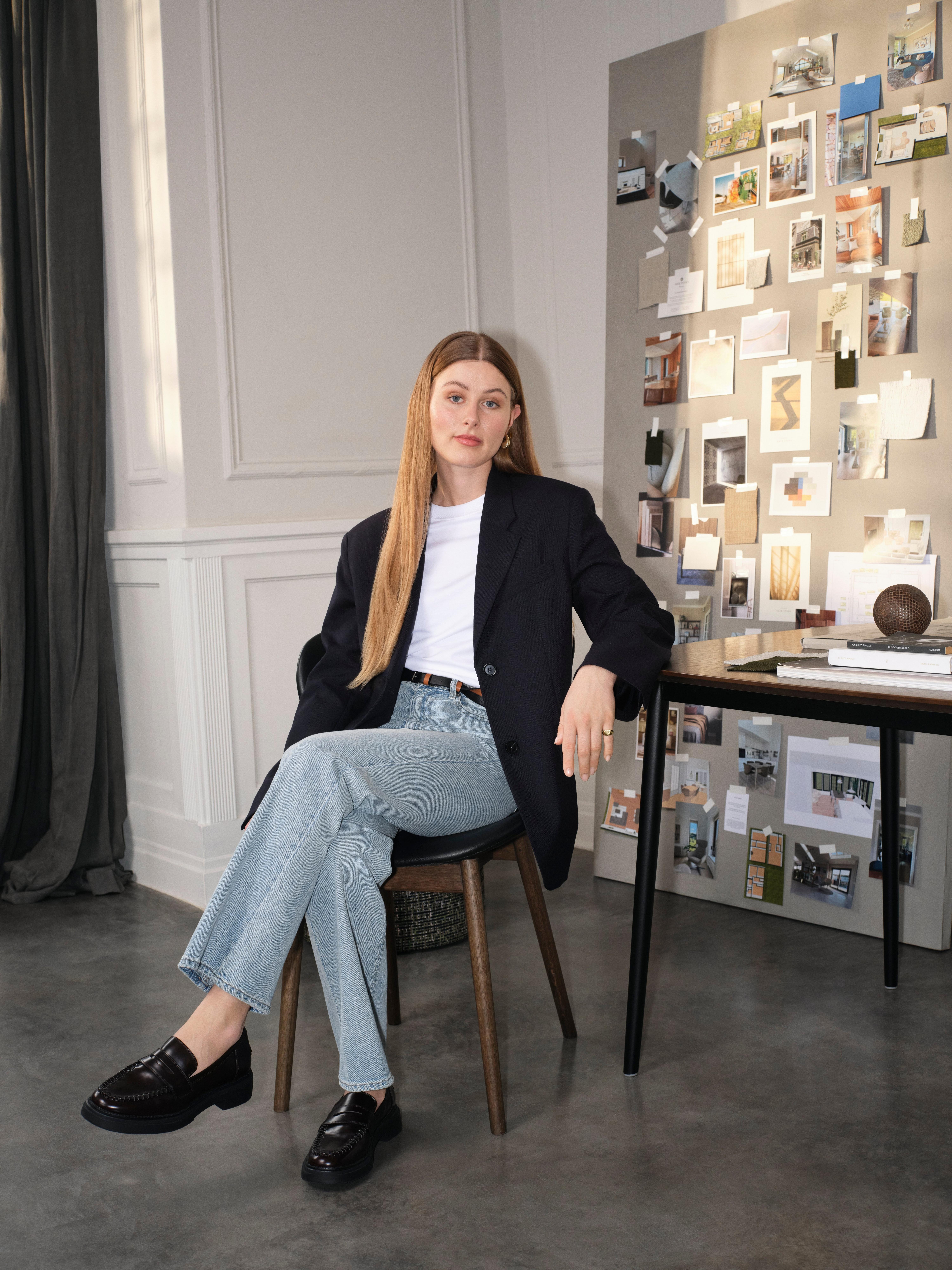 Eine Frau sitzt auf einem Stuhl, trägt Blazer und Jeans, neben einer Pinnwand voller Fotos.