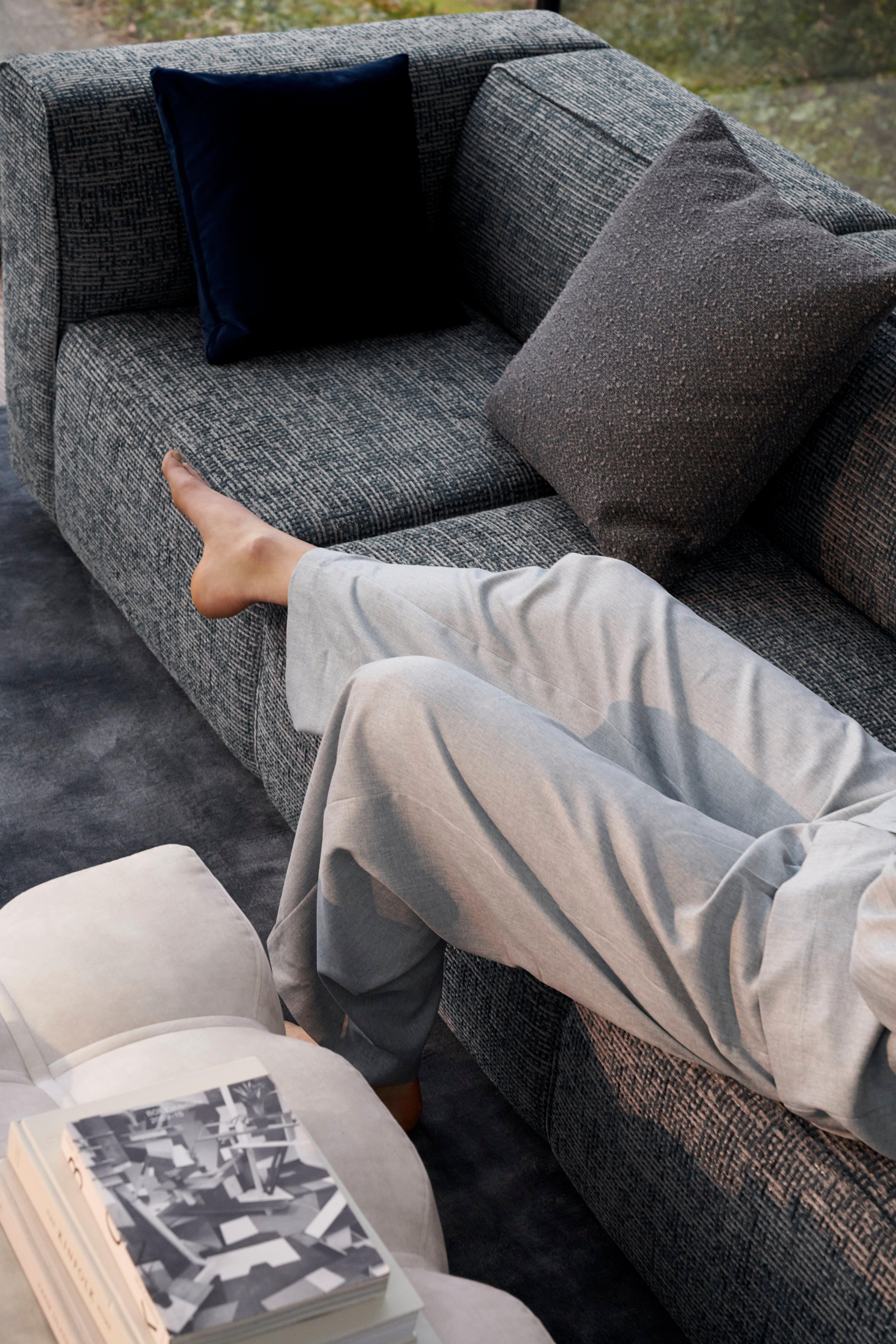 Persona con pantalones gris claro descansando en un sofá gris oscuro con cojines grises y azul marino, junto a una mesa de centro con libros.
