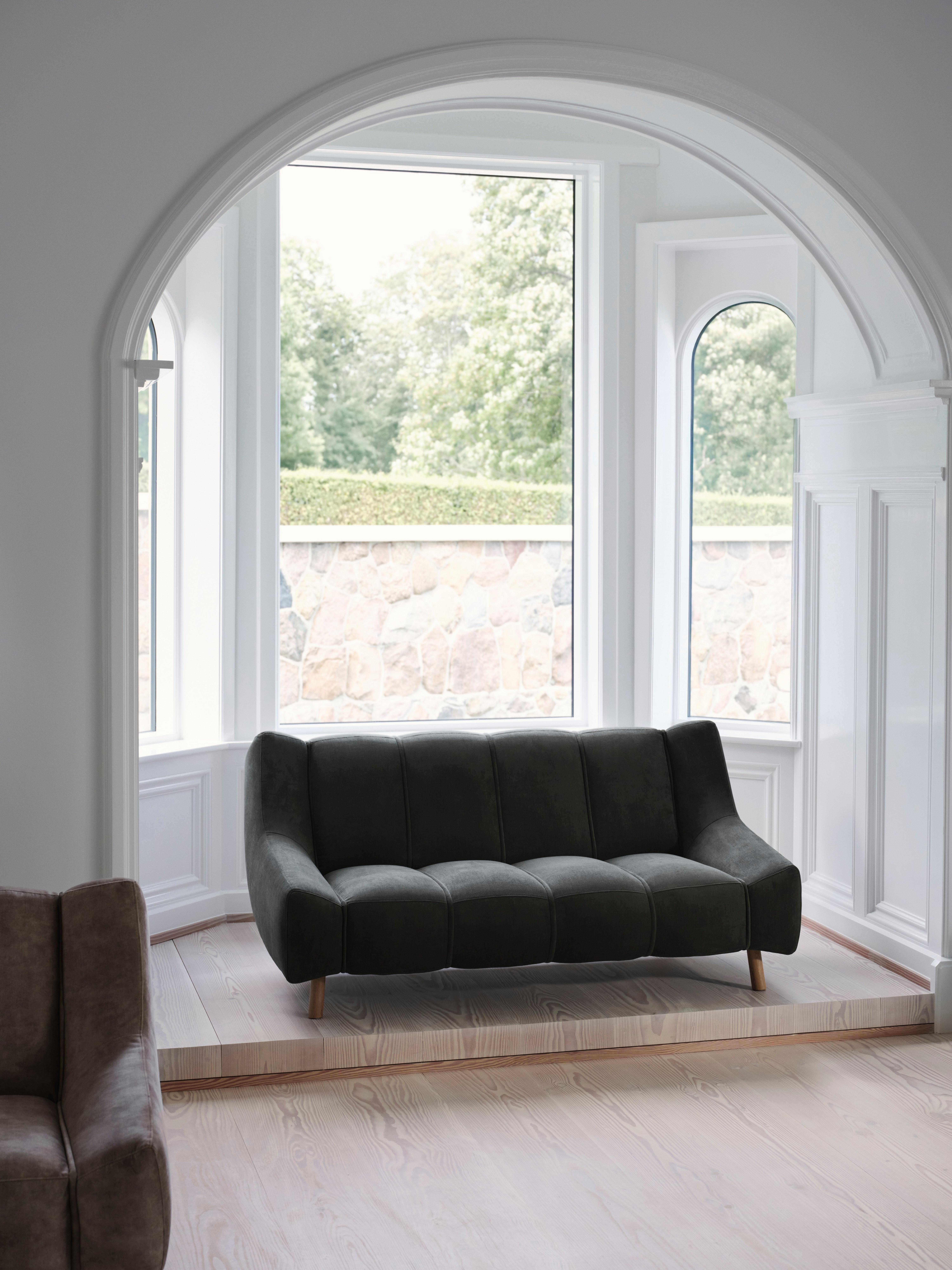 Dark green sofa on a raised wooden platform, near large windows with greenery outside.