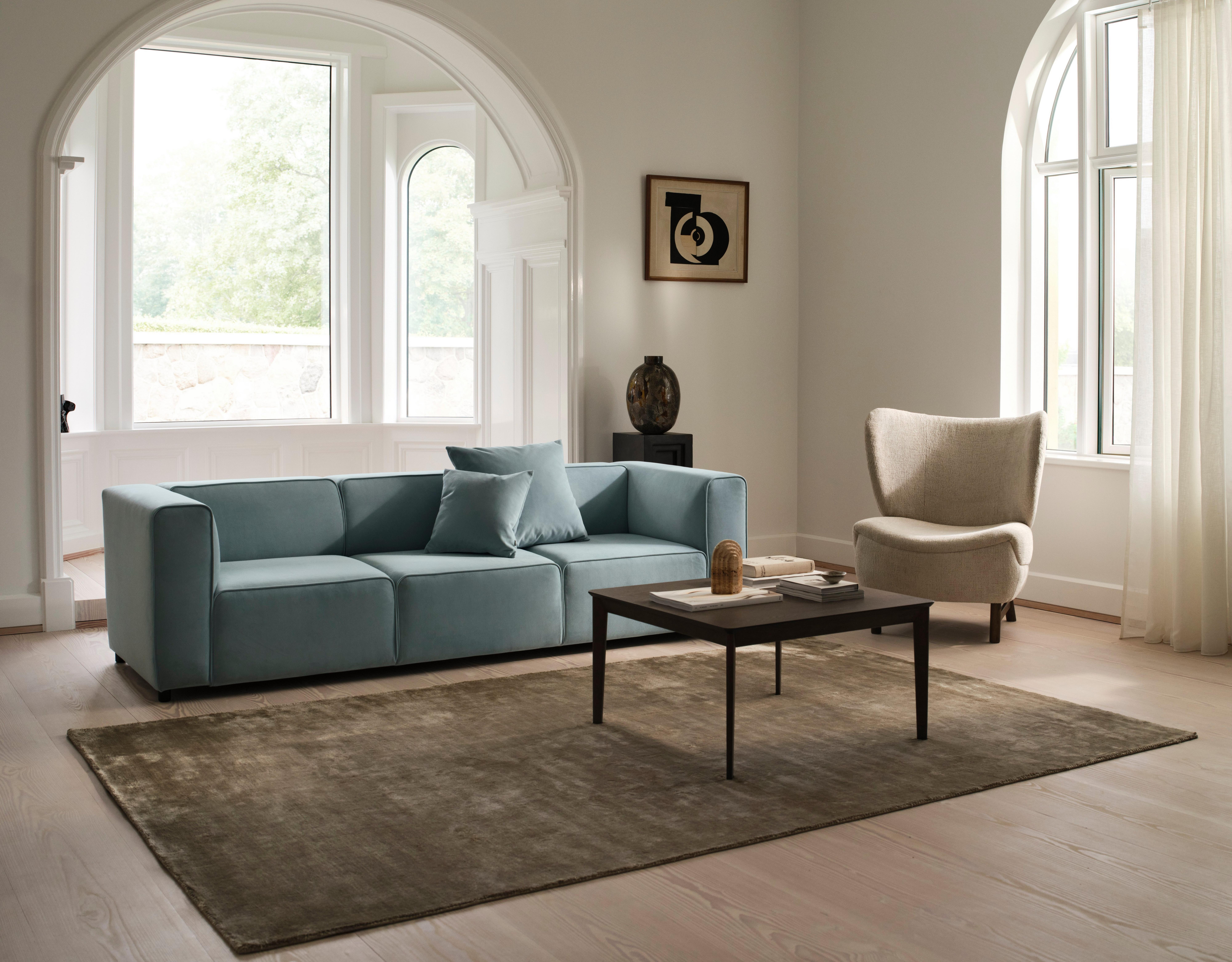 Modern living room with blue sofa, beige chair, and coffee table. Large windows and abstract wall art.