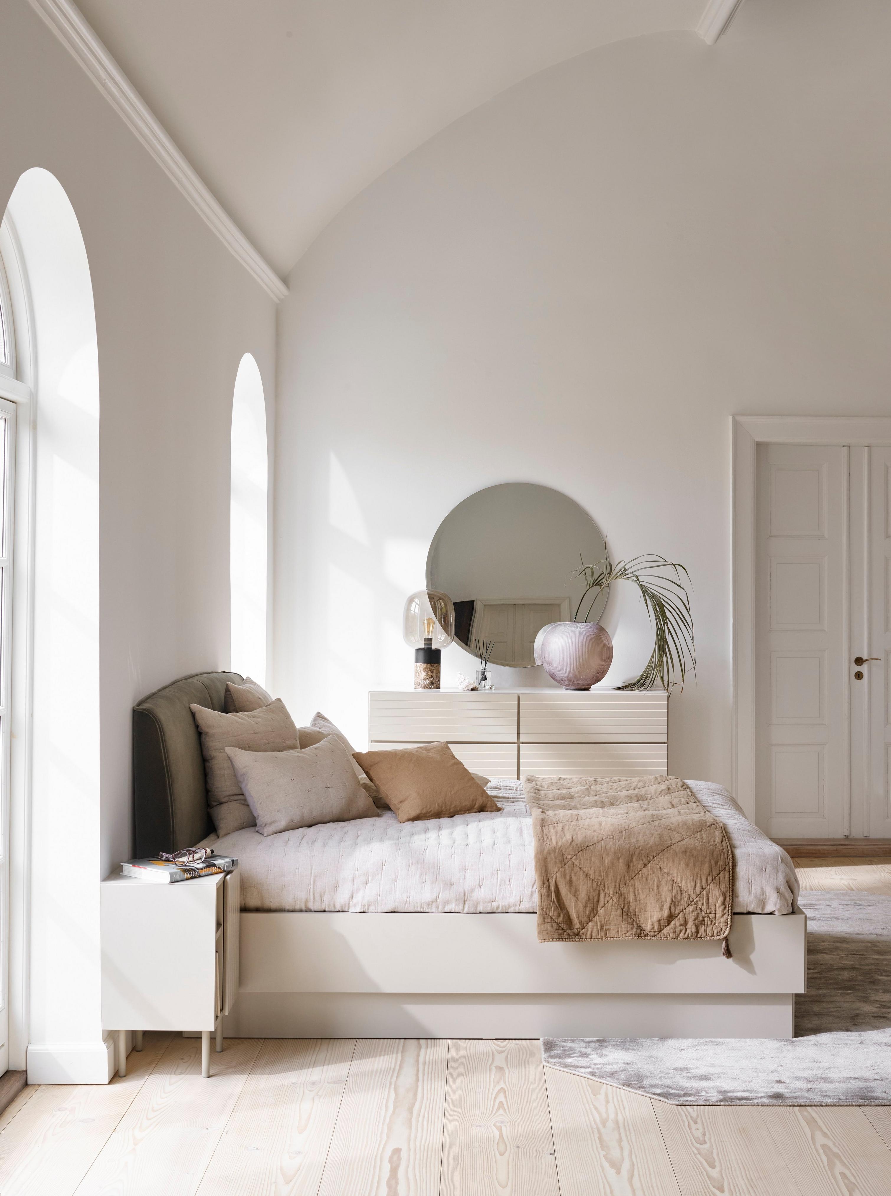 Arched bedroom with a bed, round mirror, side table, plant, and neutral-toned bedding.
