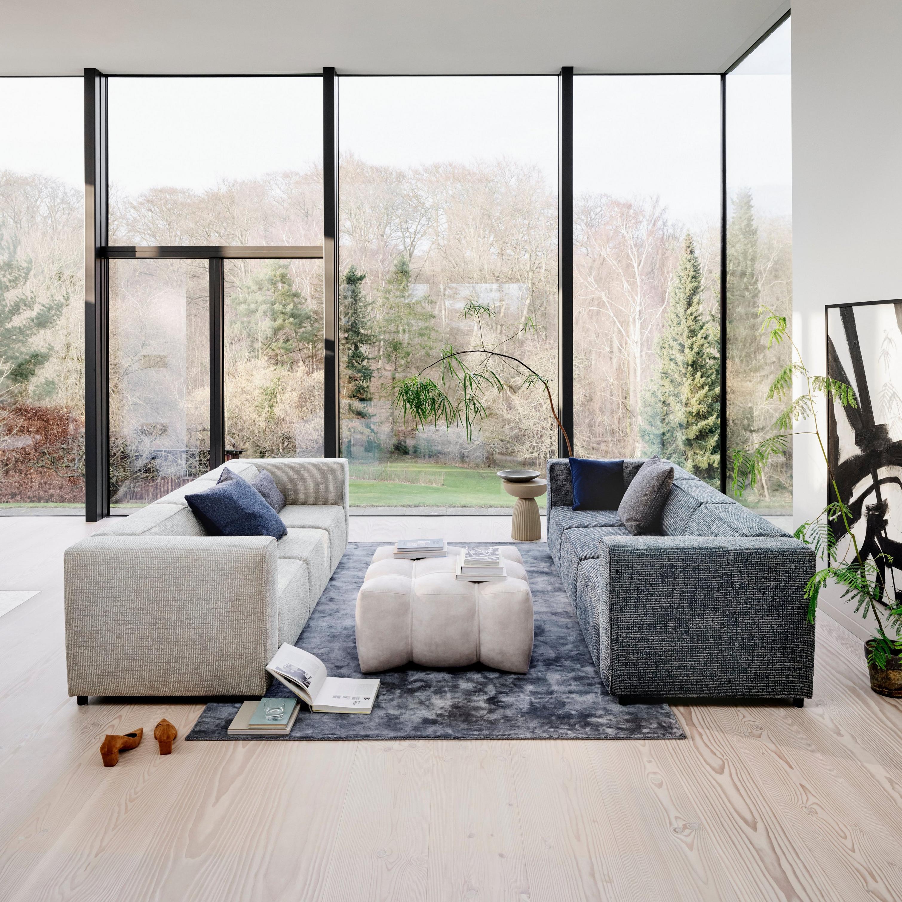 Spacious living room with two sofas facing each other, a large rug, Nawabari pouf, floor-to-ceiling windows, and a view of trees.