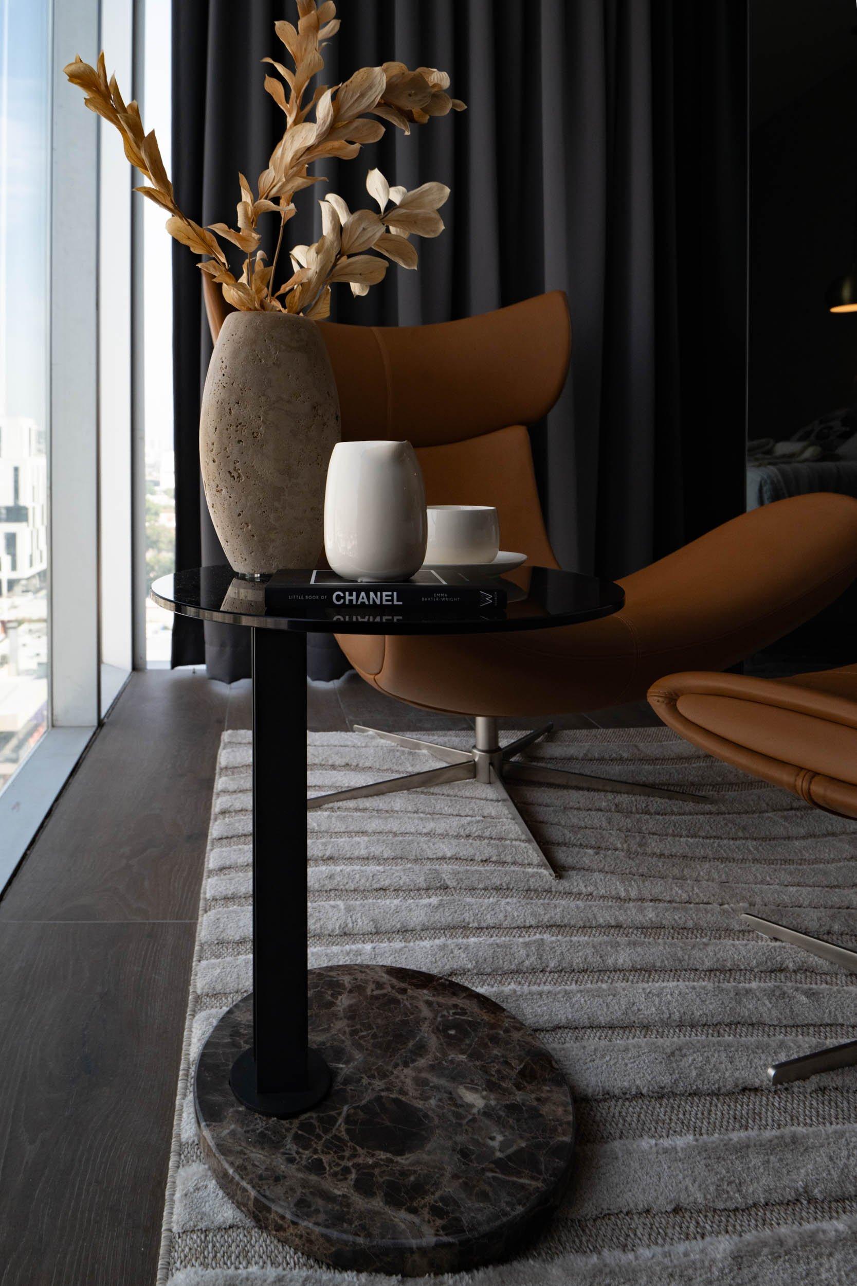 Table d’appoint ronde avec vase, tasses et livre CHANEL, près d’un fauteuil.